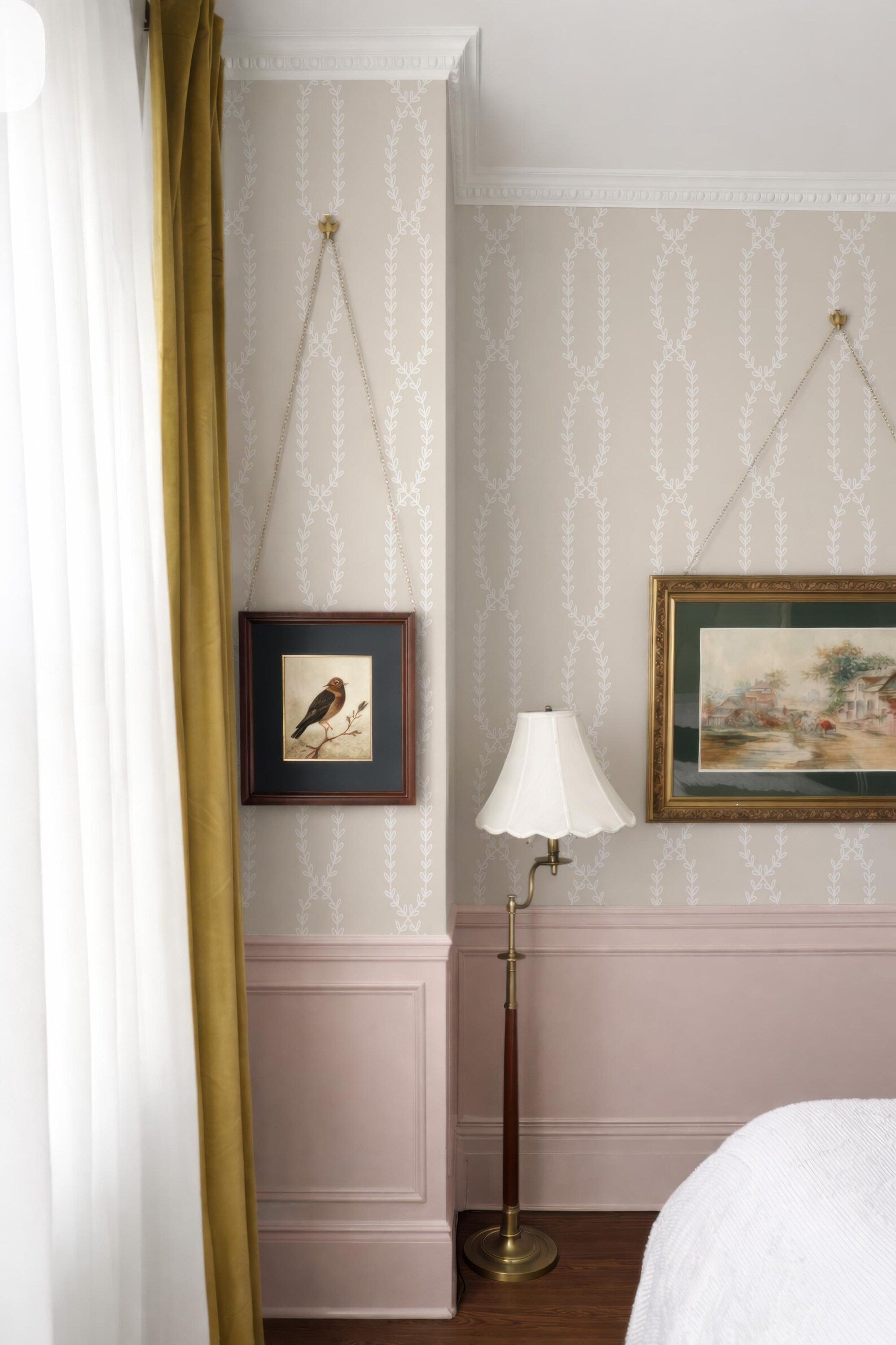 a bedroom with a bed, a lamp, and two framed pictures hanging on the wall. The room has a pink and white color scheme, and the curtains are open, allowing natural light to enter the space.