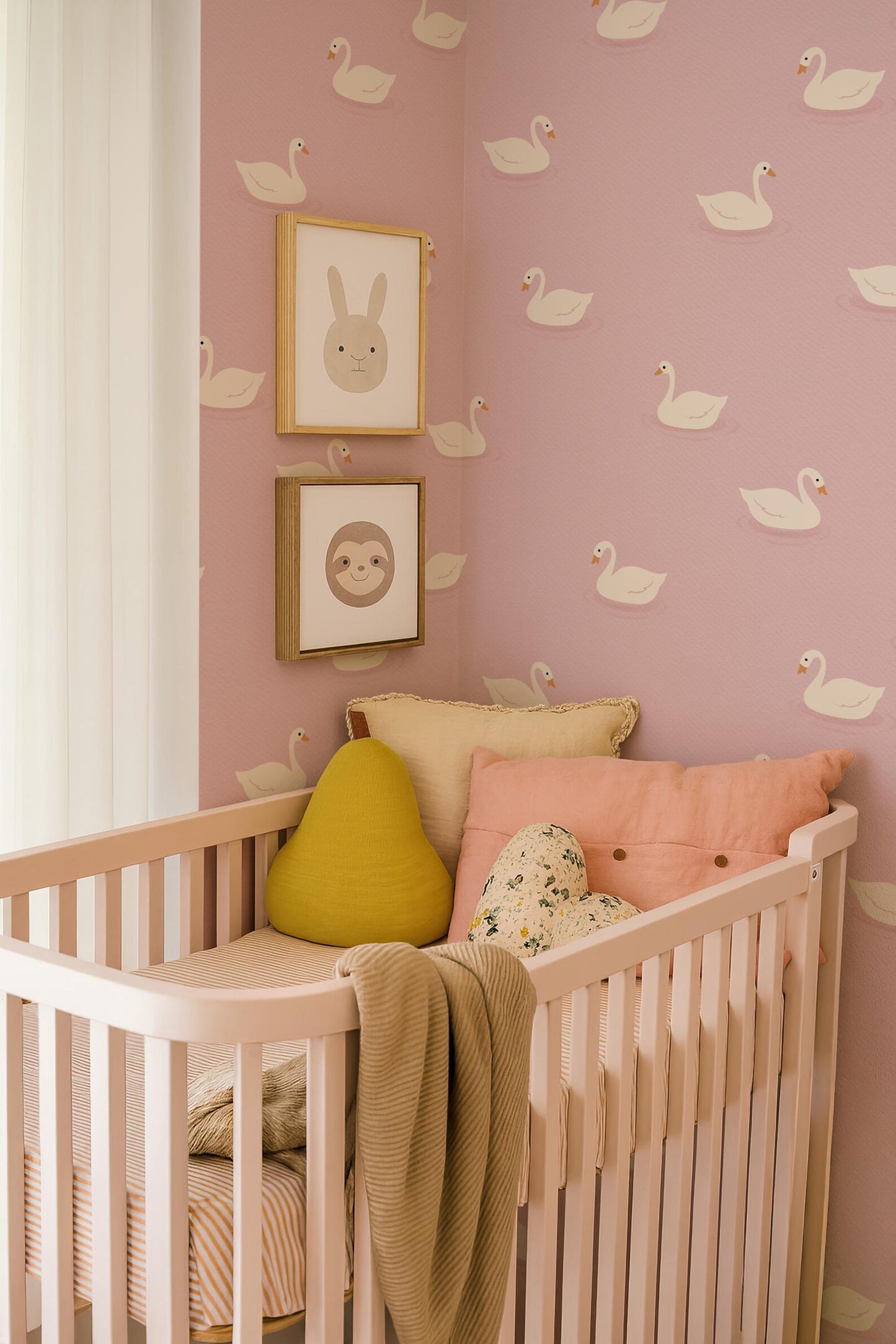 a nursery with a pink wall decorated with white swan patterns. In the corner of the room, there is a white crib with a yellow pillow and a pink blanket. Above the crib, there are two framed pictures of swan faces.