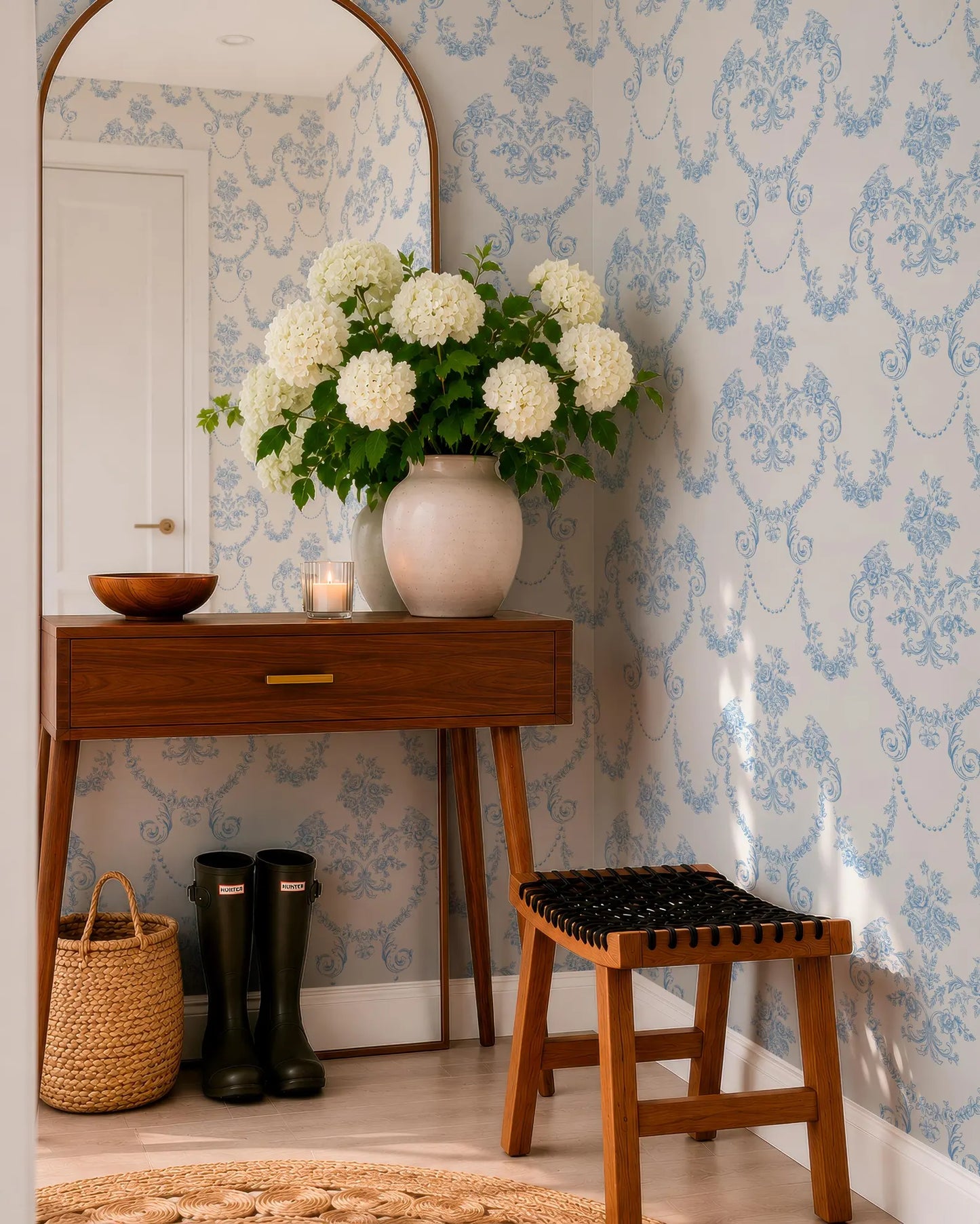 a cozy corner of a room with a wooden dresser, a mirror, a vase of white hydrangeas, a pair of black rain boots, and a black woven stool.
