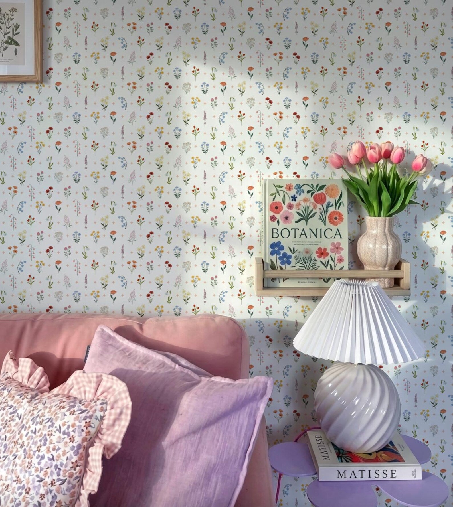 a cozy living space with a floral wallpaper, a pink sofa, a lamp, a vase of pink tulips, and a framed botanical illustration on the wall.
