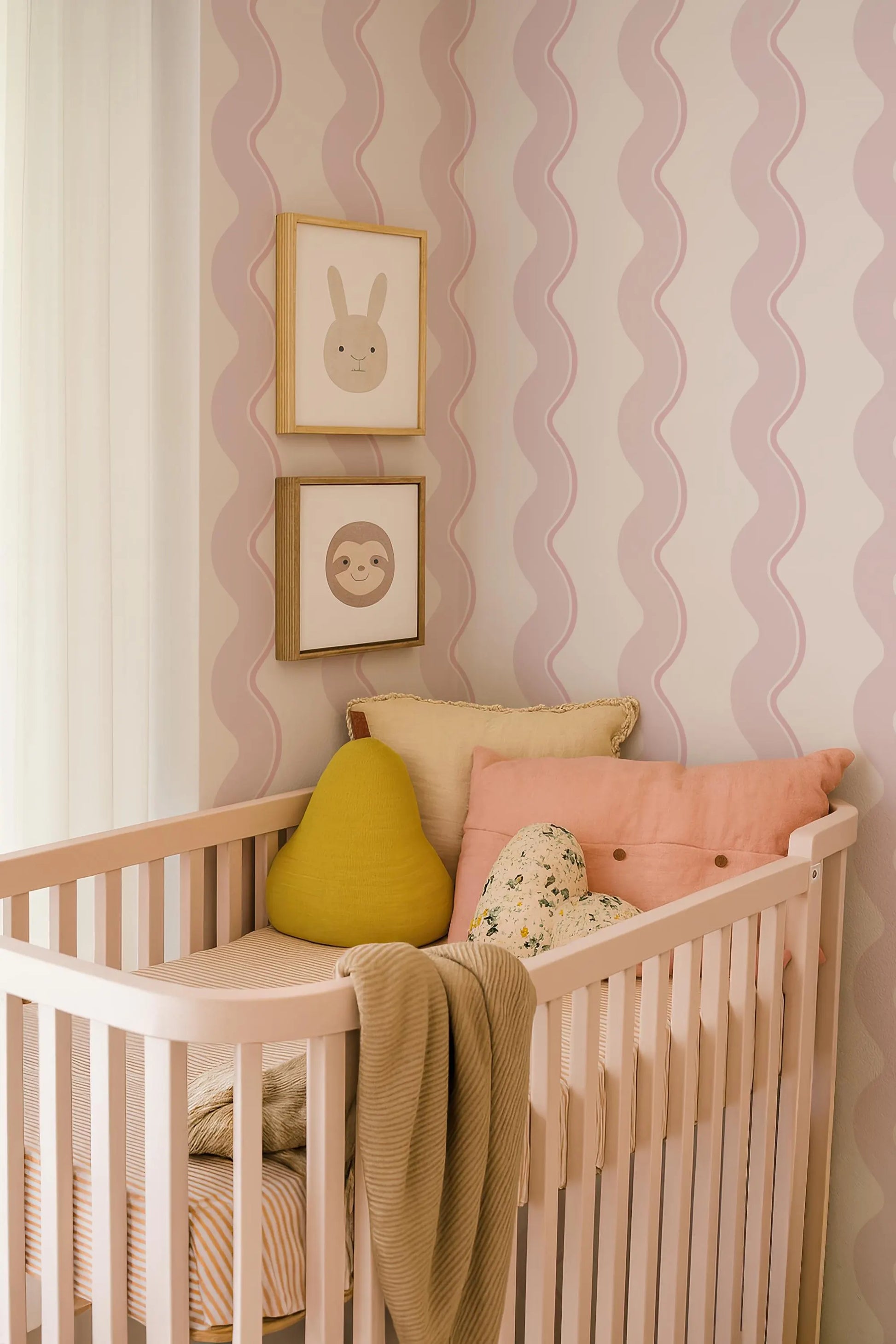a cozy nursery with a white crib, adorned with a yellow pillow, a pink pillow, and a floral pillow. The crib is situated against a wall with pink and white striped wallpaper, and there are two framed pictures hanging above it.