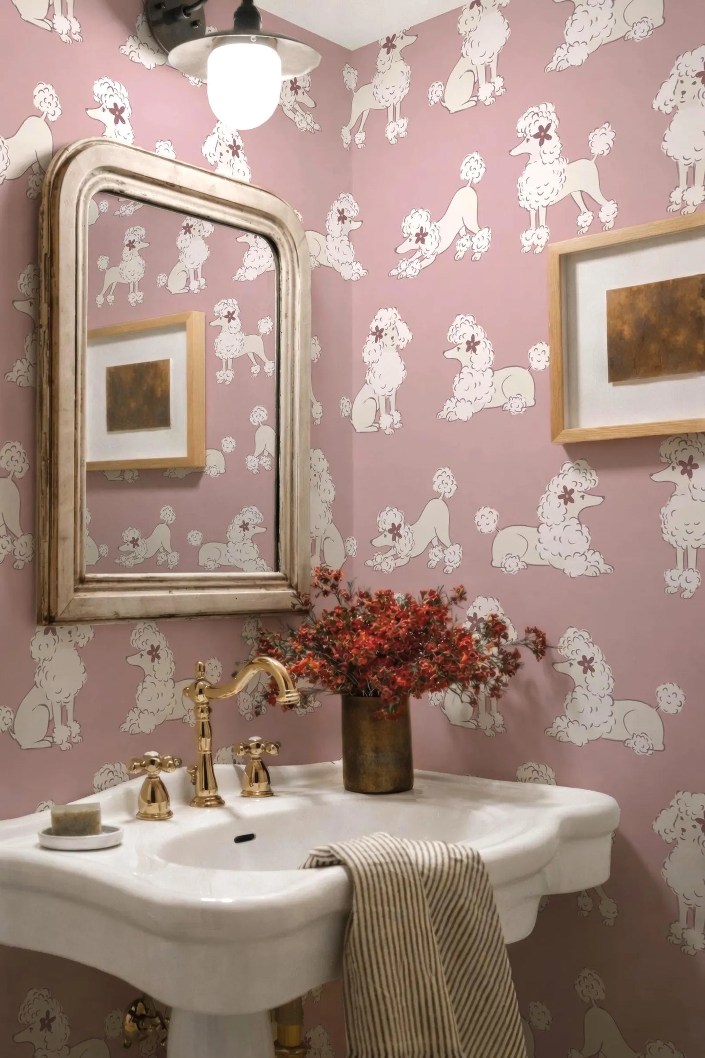 a bathroom with pink wallpaper decorated with white poodle illustrations. The bathroom has a white pedestal sink with a gold faucet, and a framed picture on the wall above the sink. There is a vase of red flowers on the sink, and a striped towel is hanging on the side.