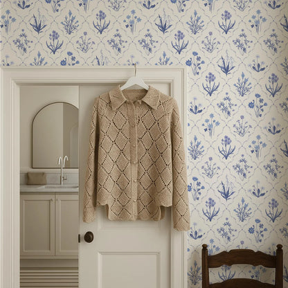 a cozy, beige knitted sweater hanging on a white door, with a blue floral wallpaper behind it. The sweater has a diamond pattern and appears to be made of a soft, warm material. The door is set in a room with a sink and a mirror, creating a homely atmosphere.