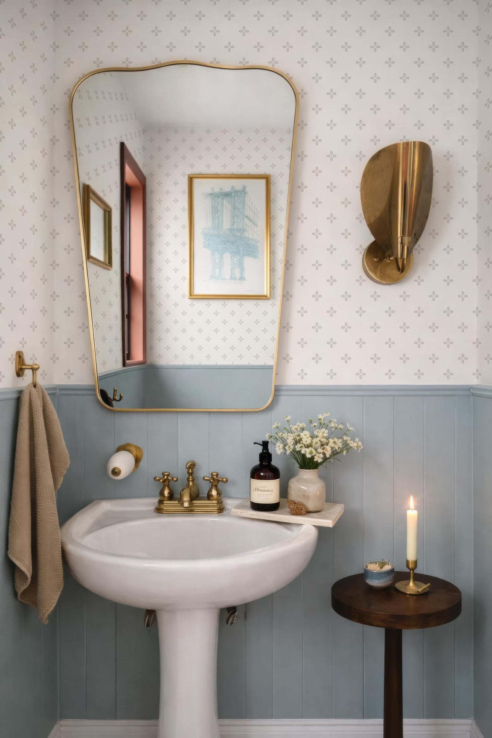 a bathroom with a white pedestal sink, a mirror, and a small table with a candle. The walls are covered in blue and white patterned wallpaper, and there is a framed picture on the wall.