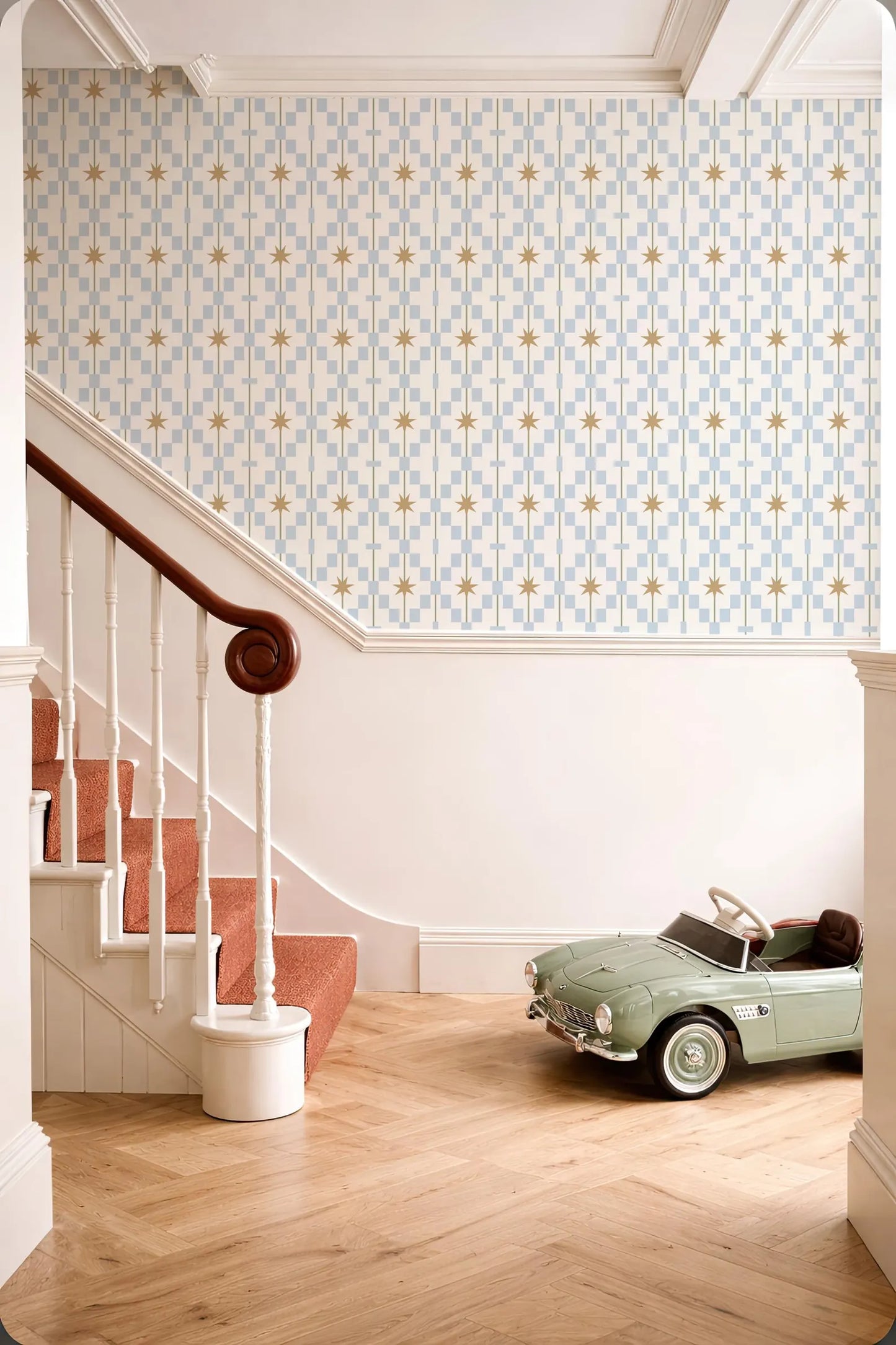 a vintage-style green toy car parked on a wooden floor next to a staircase with a white railing. The walls are adorned with a patterned wallpaper, and the overall color scheme is predominantly white and blue.