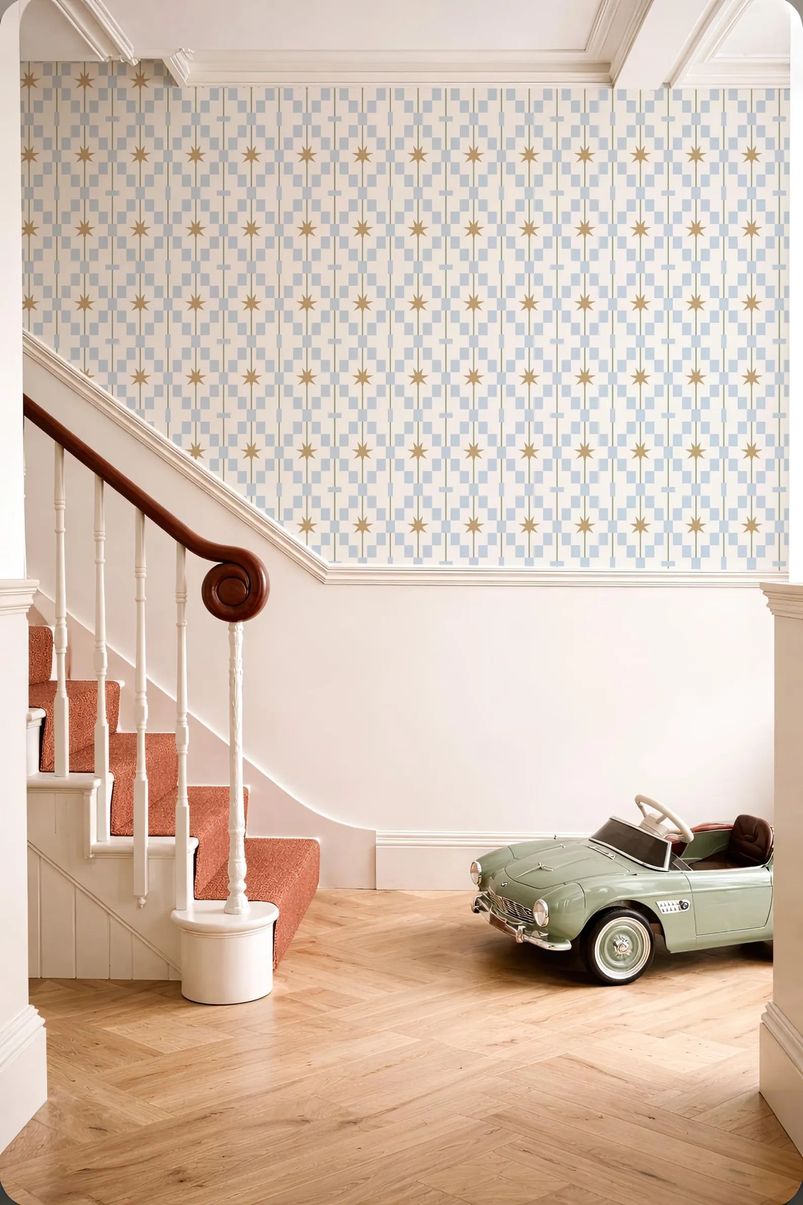 a vintage-style green toy car parked on a wooden floor next to a staircase with a white railing. The walls are adorned with a patterned wallpaper, and the overall color scheme is predominantly white and blue.