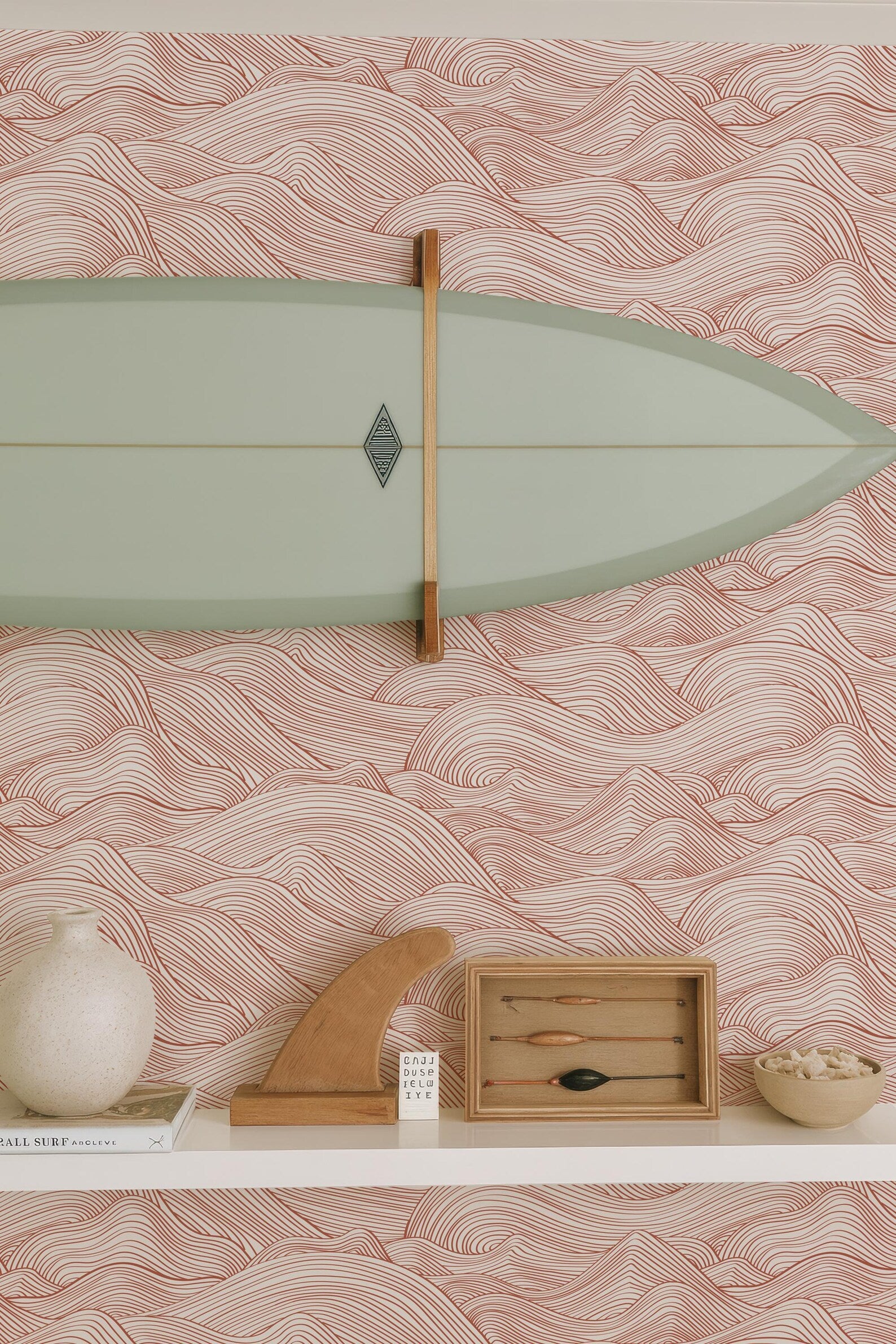 a shelf with various items, including a surfboard, a wooden sculpture, a small wooden box, and a white vase. The wall behind the shelf has a pink and white wave-patterned wallpaper.