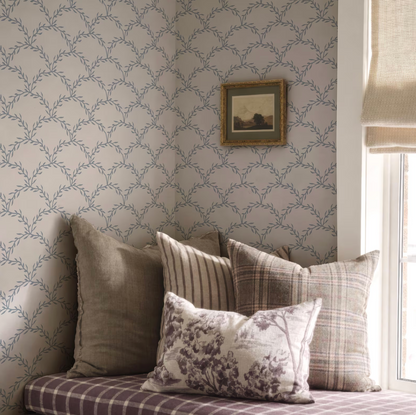 a cozy corner of a room with a patterned wallpaper, a window, and a framed picture on the wall. The room has a mix of neutral and patterned pillows on a couch, creating a comfortable and inviting atmosphere.