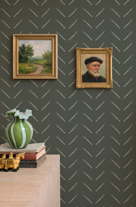 a wall with two framed paintings hanging on it, one depicting a landscape and the other a portrait of an older man. There is also a green vase and a stack of books on a wooden surface below the paintings.