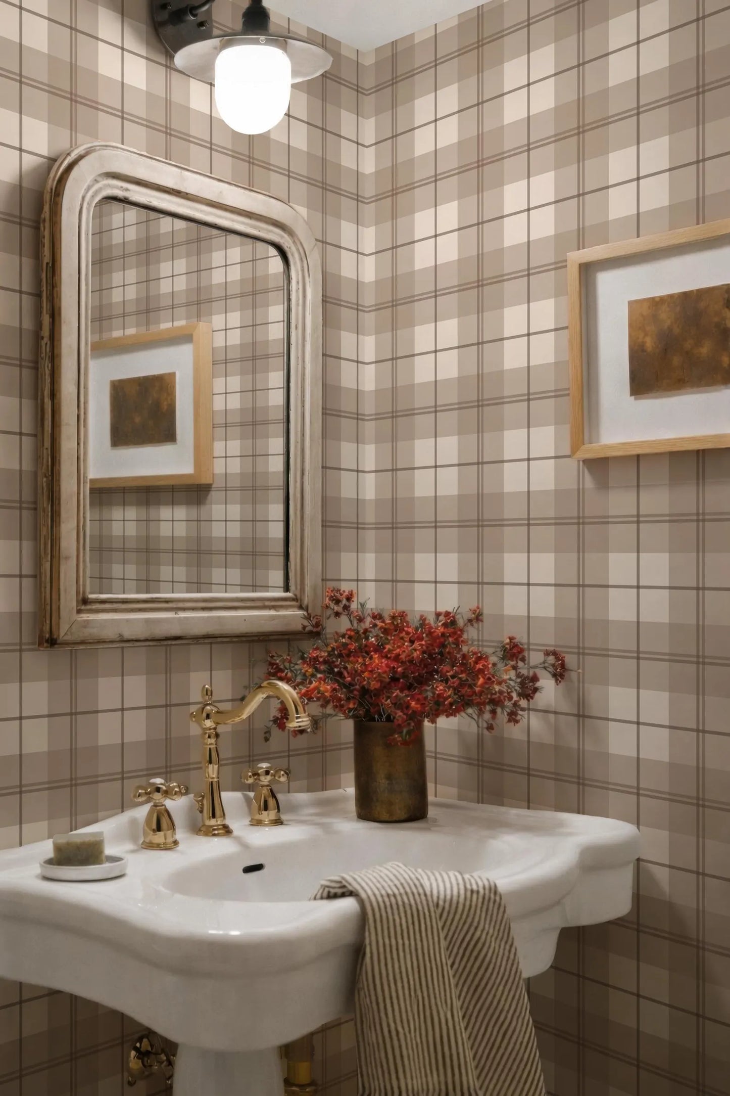 a bathroom with a white pedestal sink, a gold faucet, and a framed picture on the wall. The walls are covered in a checkered pattern, and there is a vase of red flowers on the sink.
