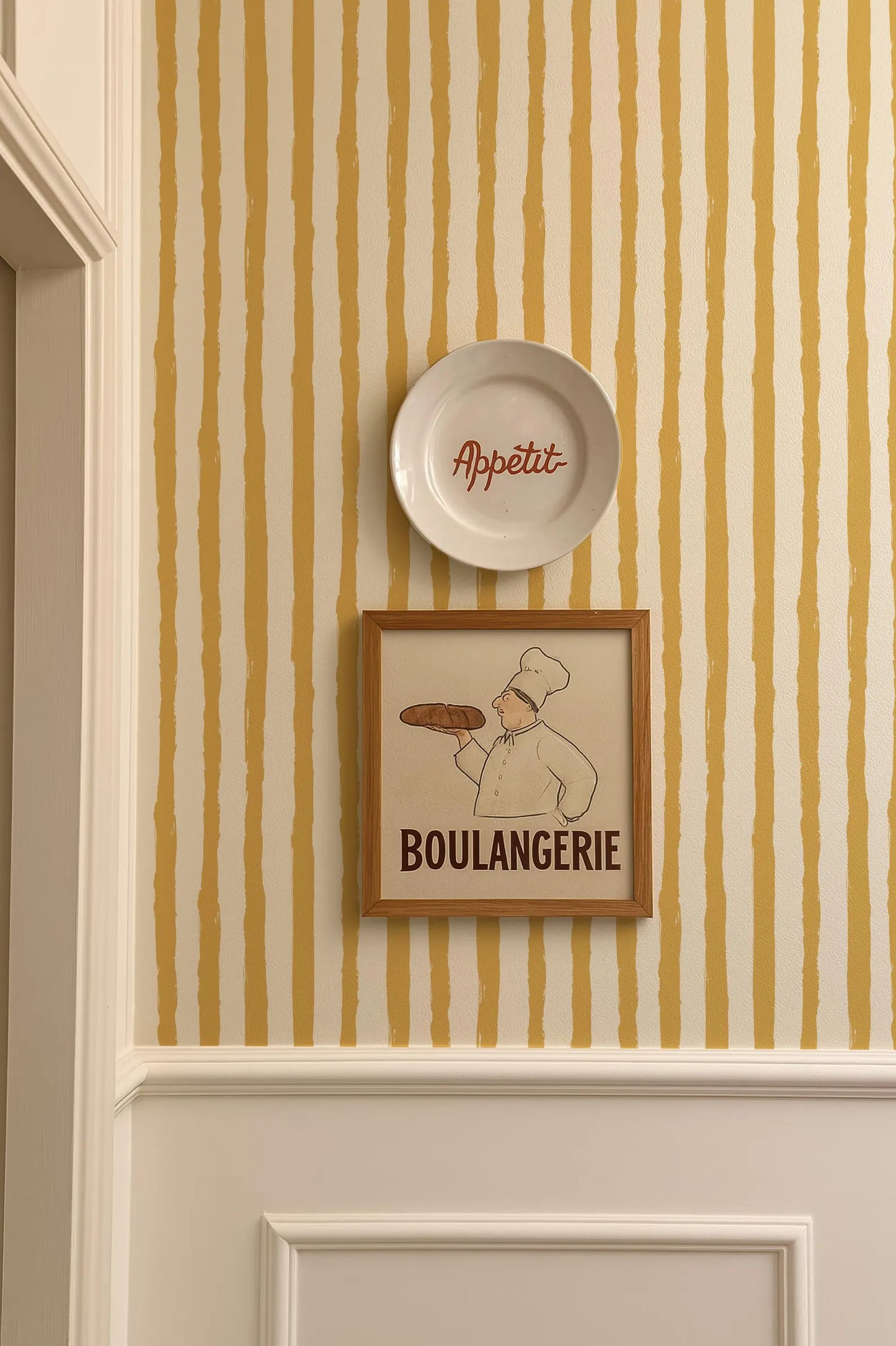 a framed illustration of a chef holding a loaf of bread, with the word "Boulangerie" written below the image. The chef is wearing a chef's hat and apron, suggesting a bakery setting. The wall behind the chef has a striped pattern in yellow and white.