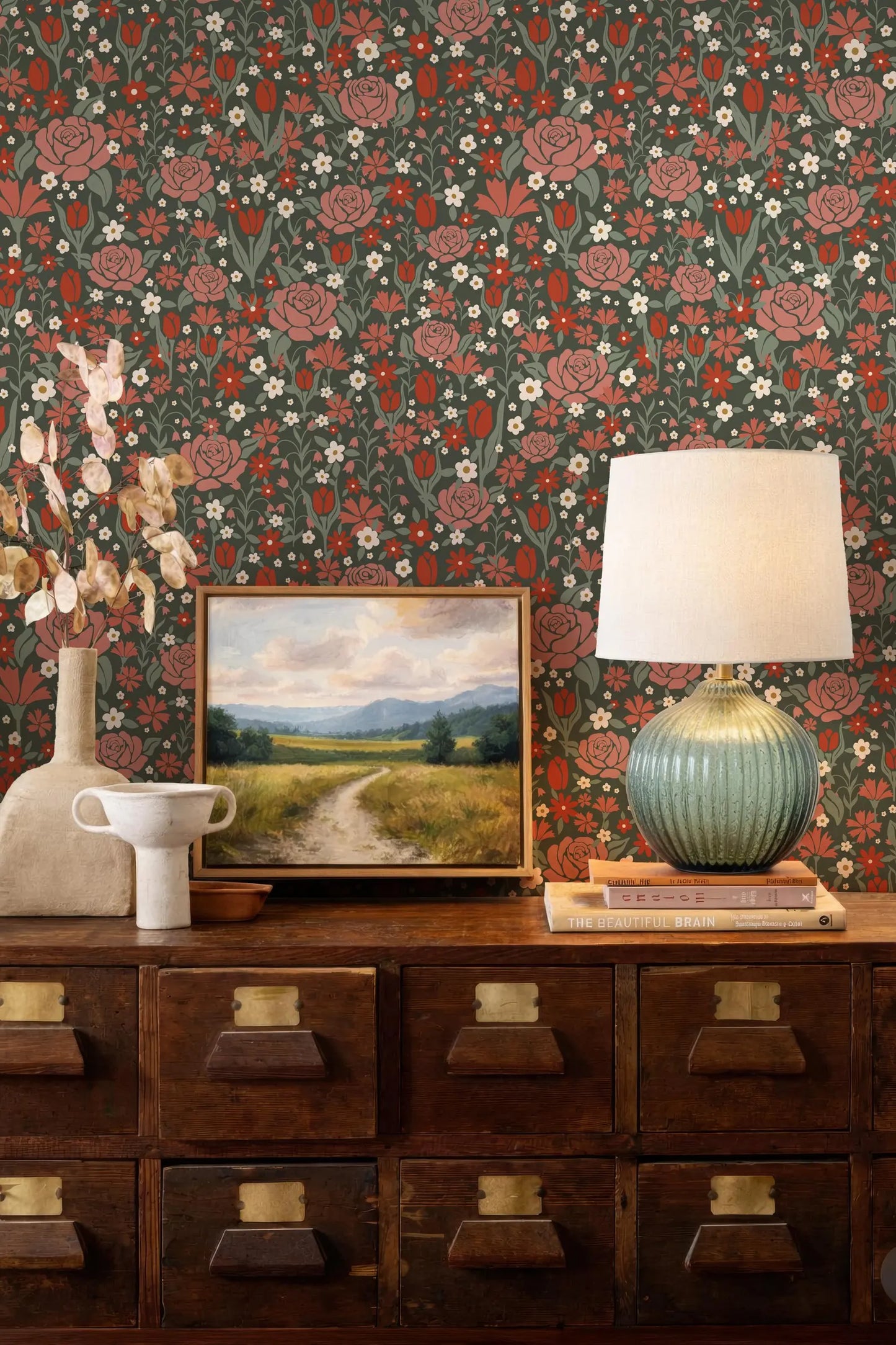 a wooden dresser with a painting, a lamp, and a vase on top. The dresser has multiple drawers and a cabinet below it. The wall behind the dresser has a floral wallpaper pattern.