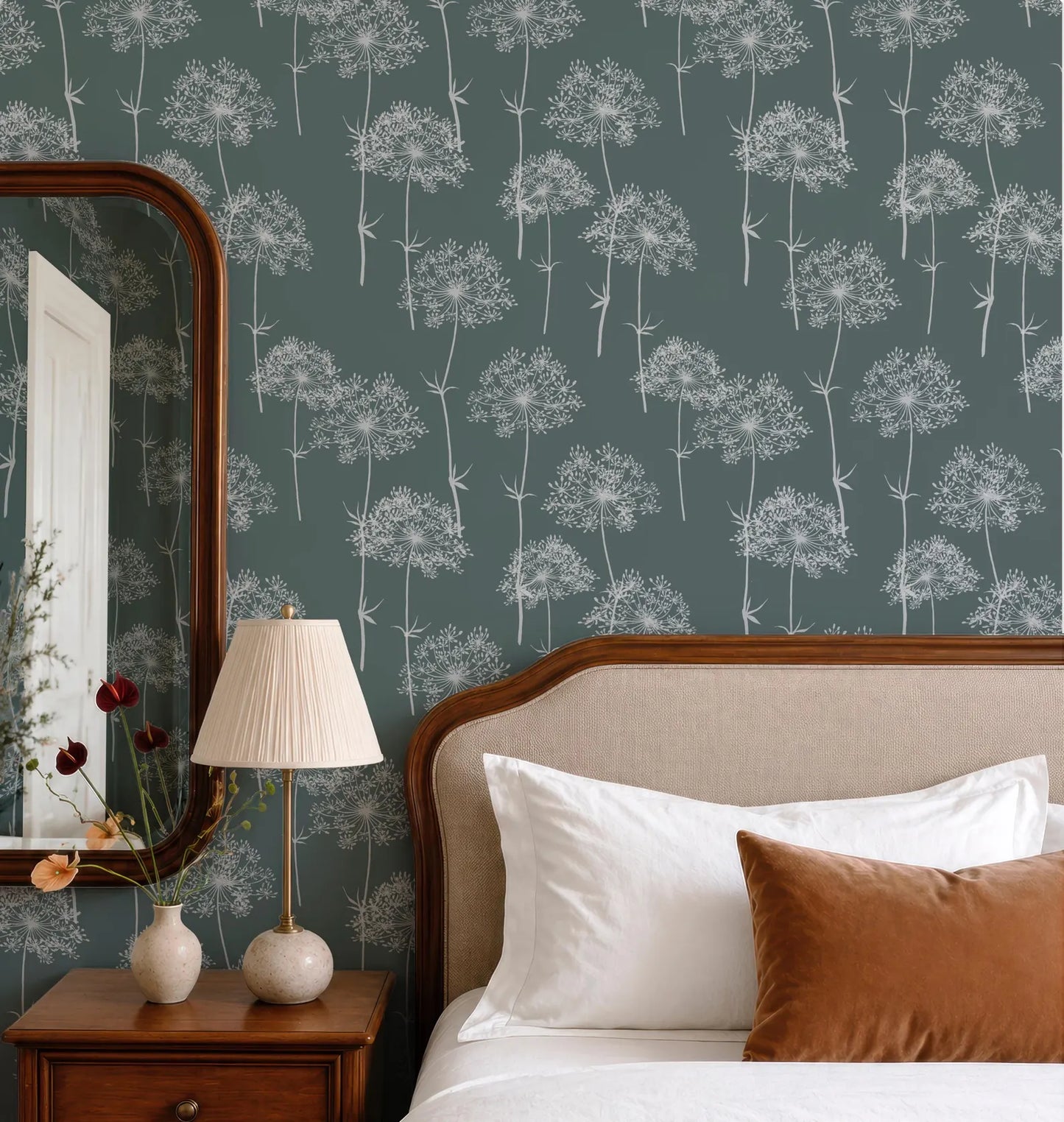 a bedroom setting with a bed, nightstand, and lamp. The bed has white pillows and a brown pillow, and the wall behind the bed has a patterned wallpaper with white flowers. The nightstand has a lamp on it, and there is a vase with flowers on the nightstand.