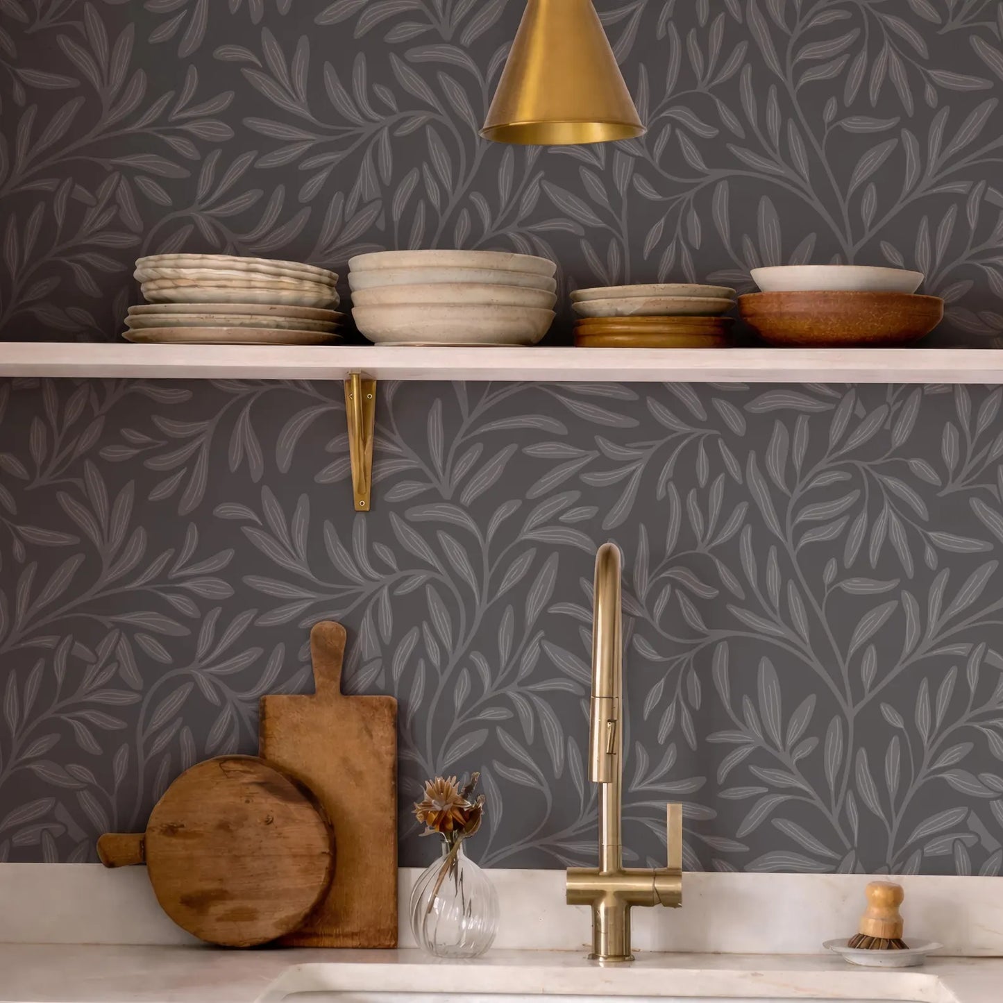 a kitchen counter with various kitchen items, including a sink, wooden cutting board, bowls, plates, and a vase with flowers. The counter has a gray and white leaf-patterned wallpaper, and a gold pendant light hangs above the counter.