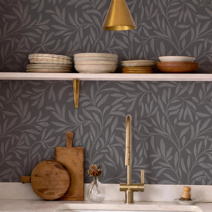 a kitchen counter with various kitchen items, including a sink, wooden cutting board, bowls, plates, and a vase with flowers. The counter has a gray and white leaf-patterned wallpaper, and a gold pendant light hangs above the counter.