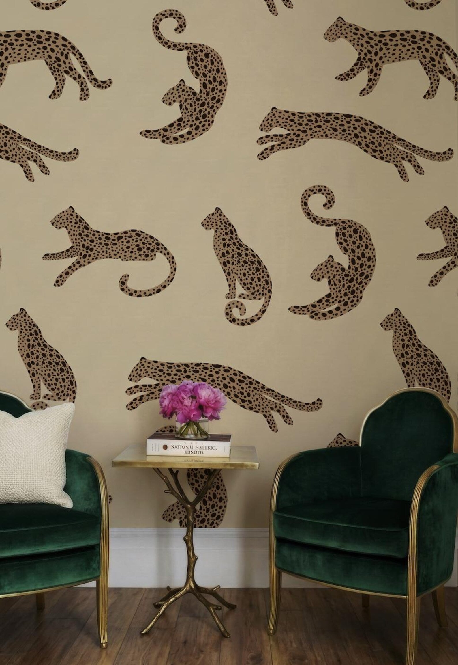 a wallpapered wall with a repeating pattern of leopard designs, including both standing and sitting leopards. In the foreground, there is a small table with a vase of pink flowers and a book, accompanied by two green velvet armchairs.