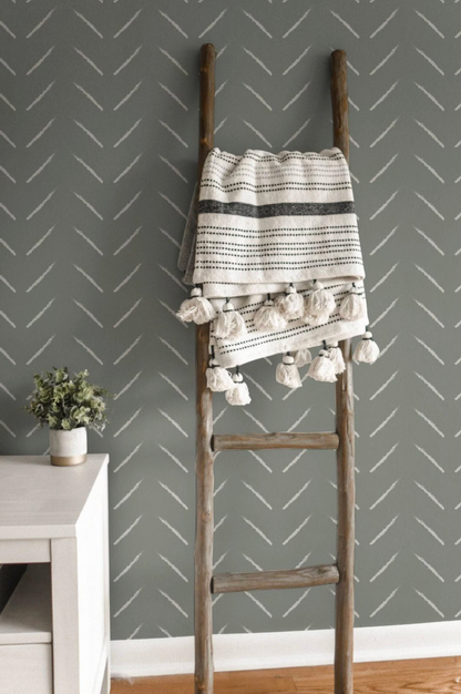 a wooden ladder with a striped blanket hanging from it, placed against a gray wall with a chevron pattern. Next to the ladder is a small potted plant, adding a touch of greenery to the scene.