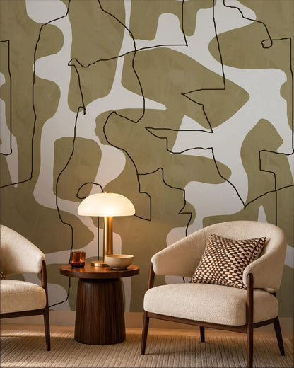 a modern living room with a minimalist design. The focal point is a wooden side table with a lamp, accompanied by two beige armchairs with patterned pillows. The wall behind the furniture is adorned with abstract art in shades of green and white, creating a visually striking backdrop.