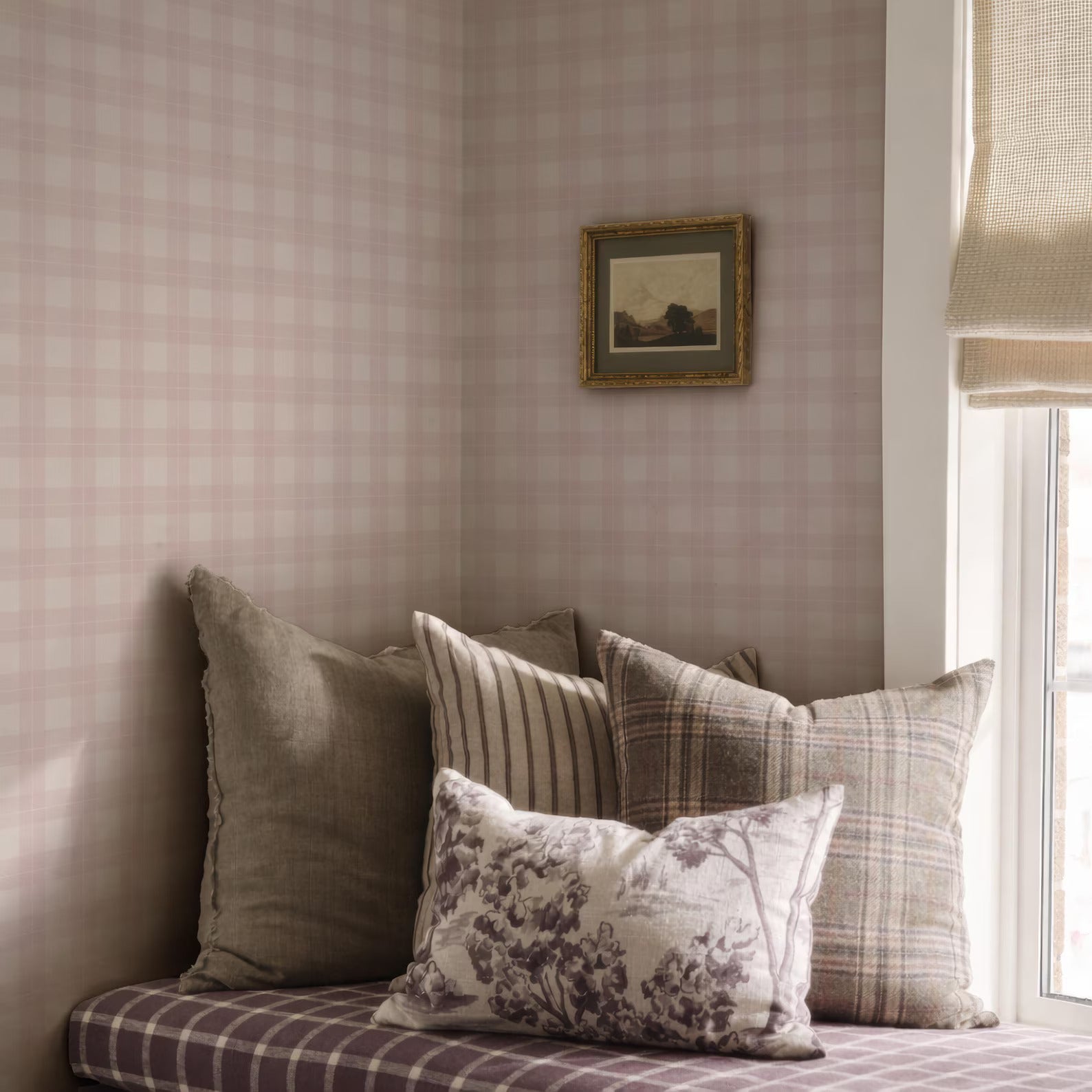 a cozy corner of a room with a plaid pattern on the walls and a window with a white curtain. The room has a comfortable seating area with a bench and several pillows, including a floral patterned pillow. A framed picture is hanging on the wall above the seating area.