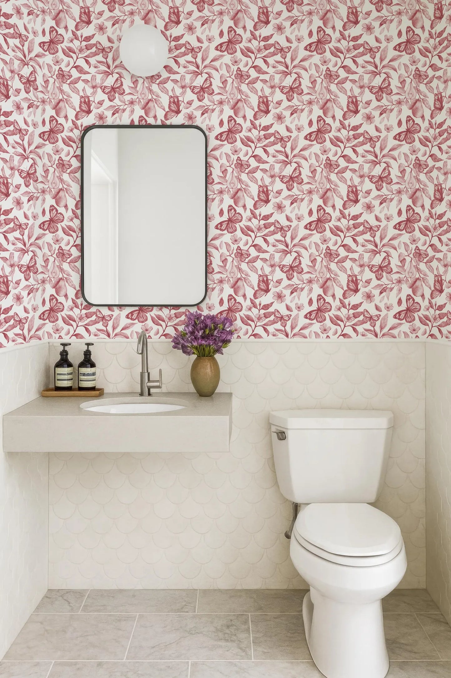 a bathroom with a white toilet, a sink with a mirror above it, and a vase of purple flowers on the counter. The walls are covered in a pink and white floral wallpaper, and the floor is tiled.