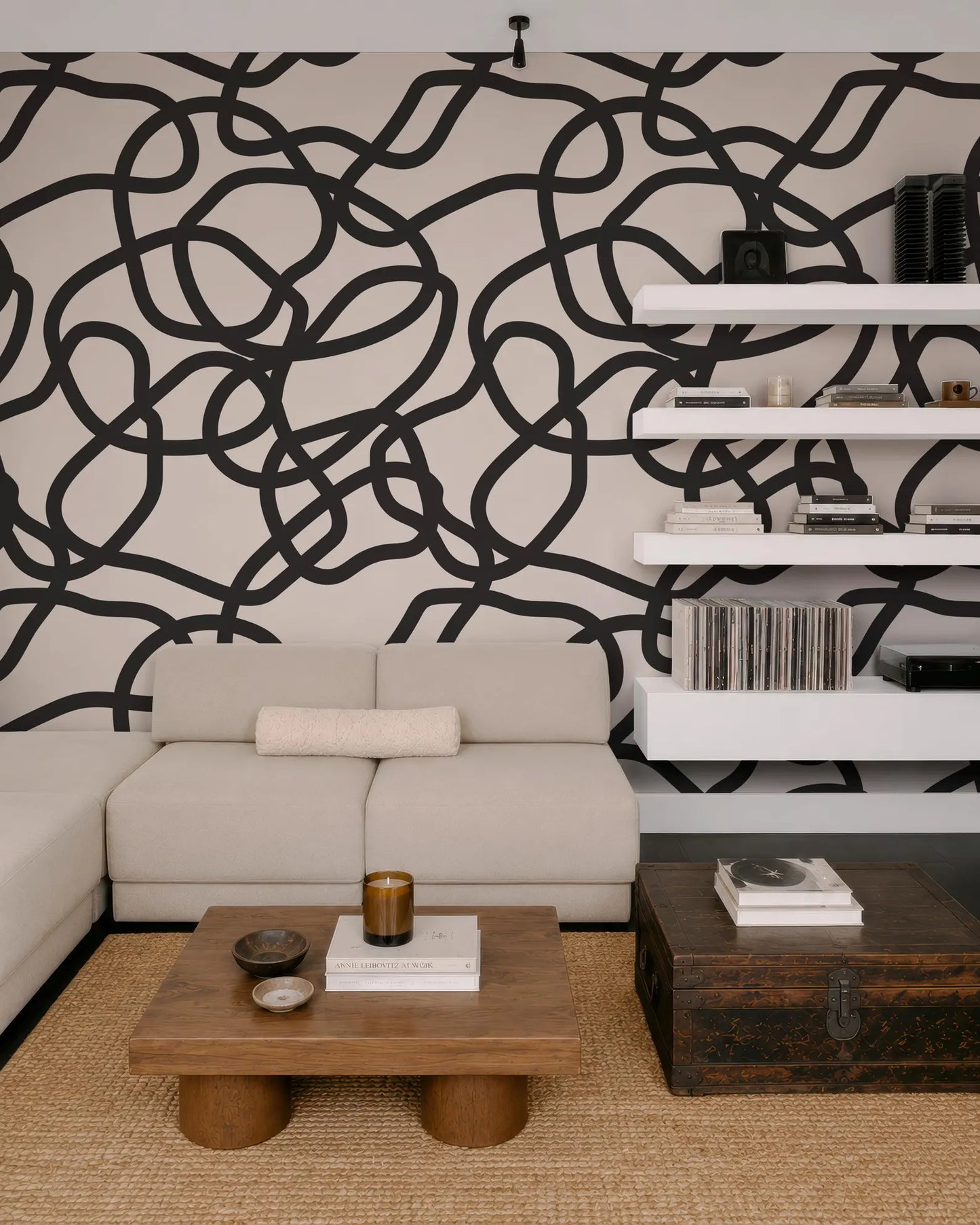 a modern living room with a large abstract black and white pattern on the wall behind a white sofa. The room is furnished with a wooden coffee table, a wooden side table, and a wooden chest. The overall design and color scheme create a contemporary and stylish atmosphere.