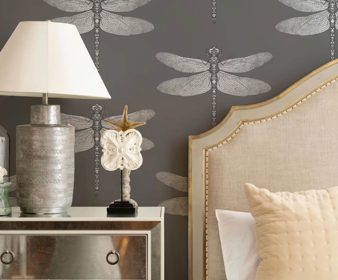 a bedroom with a gray and white dragonfly wallpaper, a white lamp, a silver and white decorative piece, and a white headboard with gold trim.