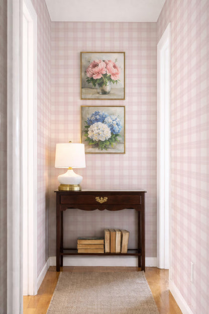 a cozy corner of a room with a wooden table, a lamp, and two framed paintings of flowers on the wall. The table has a few books on it, adding a touch of personalization to the space. The overall atmosphere of the room is warm and inviting, with the pink and white checkered wallpaper and the wooden floor contributing to the charming ambiance.