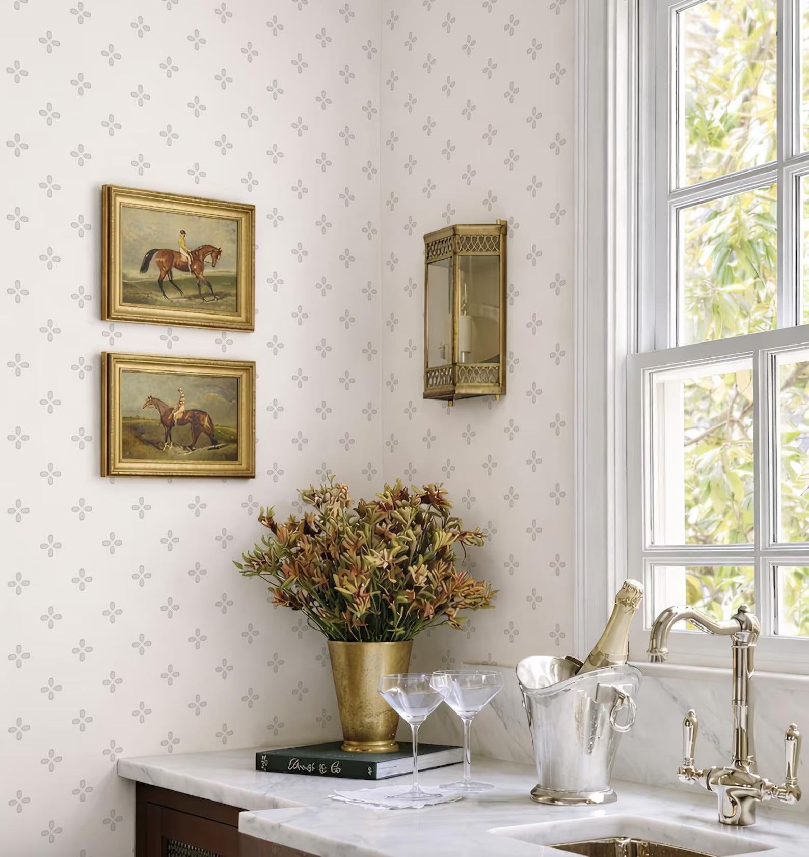 a cozy and elegant kitchen corner with a white marble countertop, a vase of flowers, a wine glass holder, and a book. The wall behind the countertop has a patterned wallpaper, and there are two framed paintings hanging on the wall.