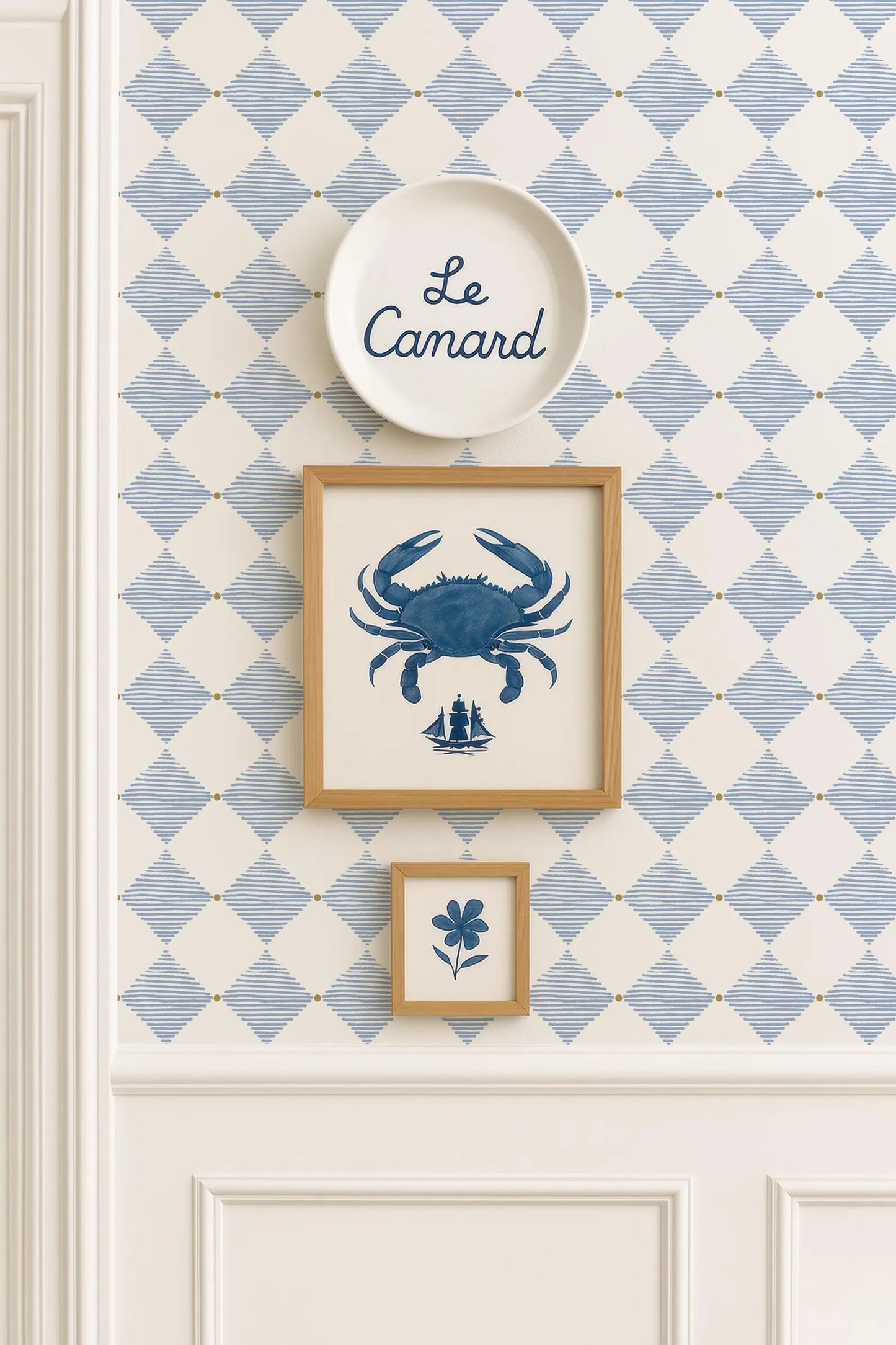 a wall with a blue and white diamond pattern wallpaper, and there are three framed pictures hanging on it. The top picture is a blue crab, the middle picture is a small blue flower, and the bottom picture is a blue and white diamond pattern.