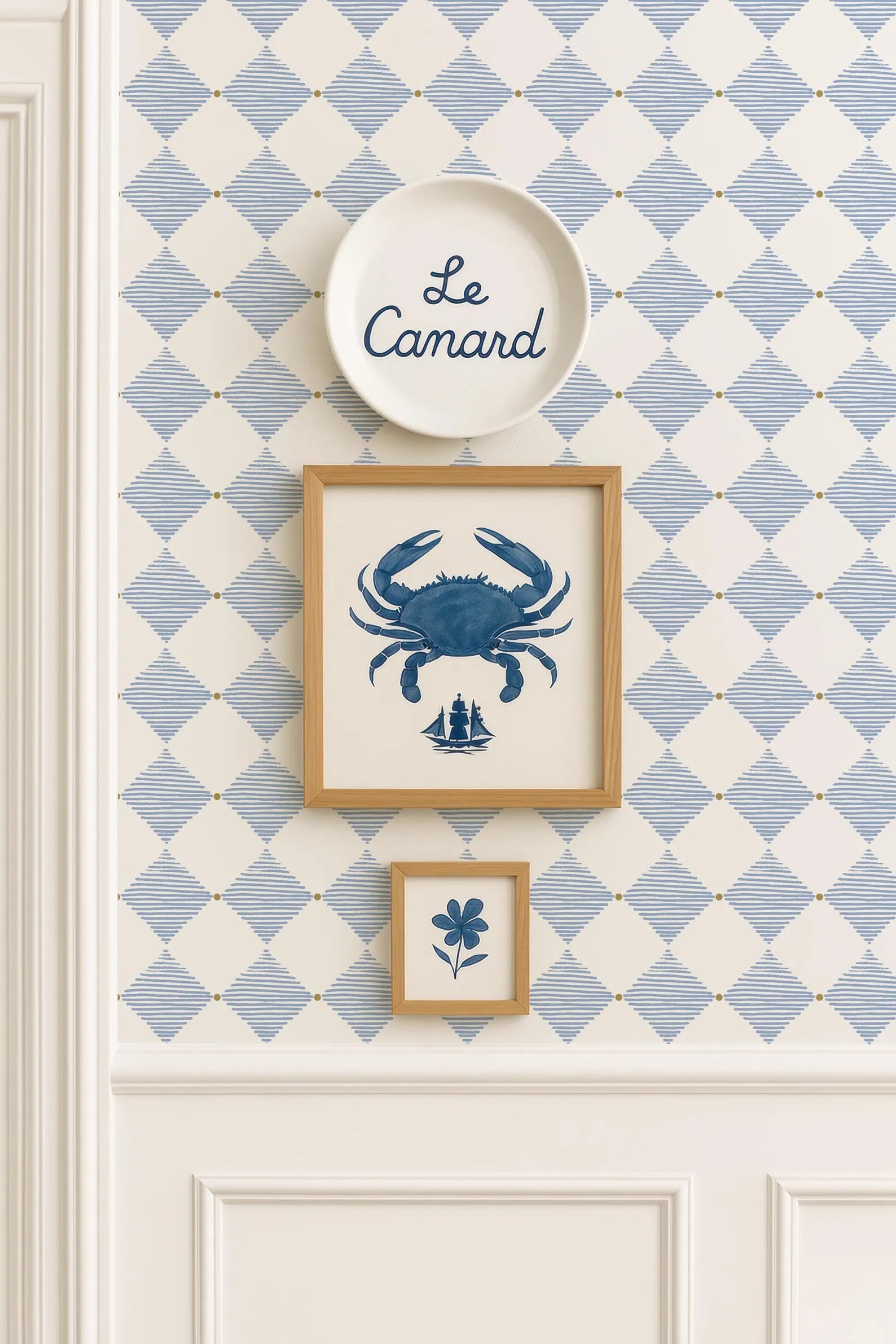 a wall with a blue and white diamond pattern wallpaper, and there are three framed pictures hanging on it. The top picture is a blue crab, the middle picture is a small blue flower, and the bottom picture is a blue and white diamond pattern.