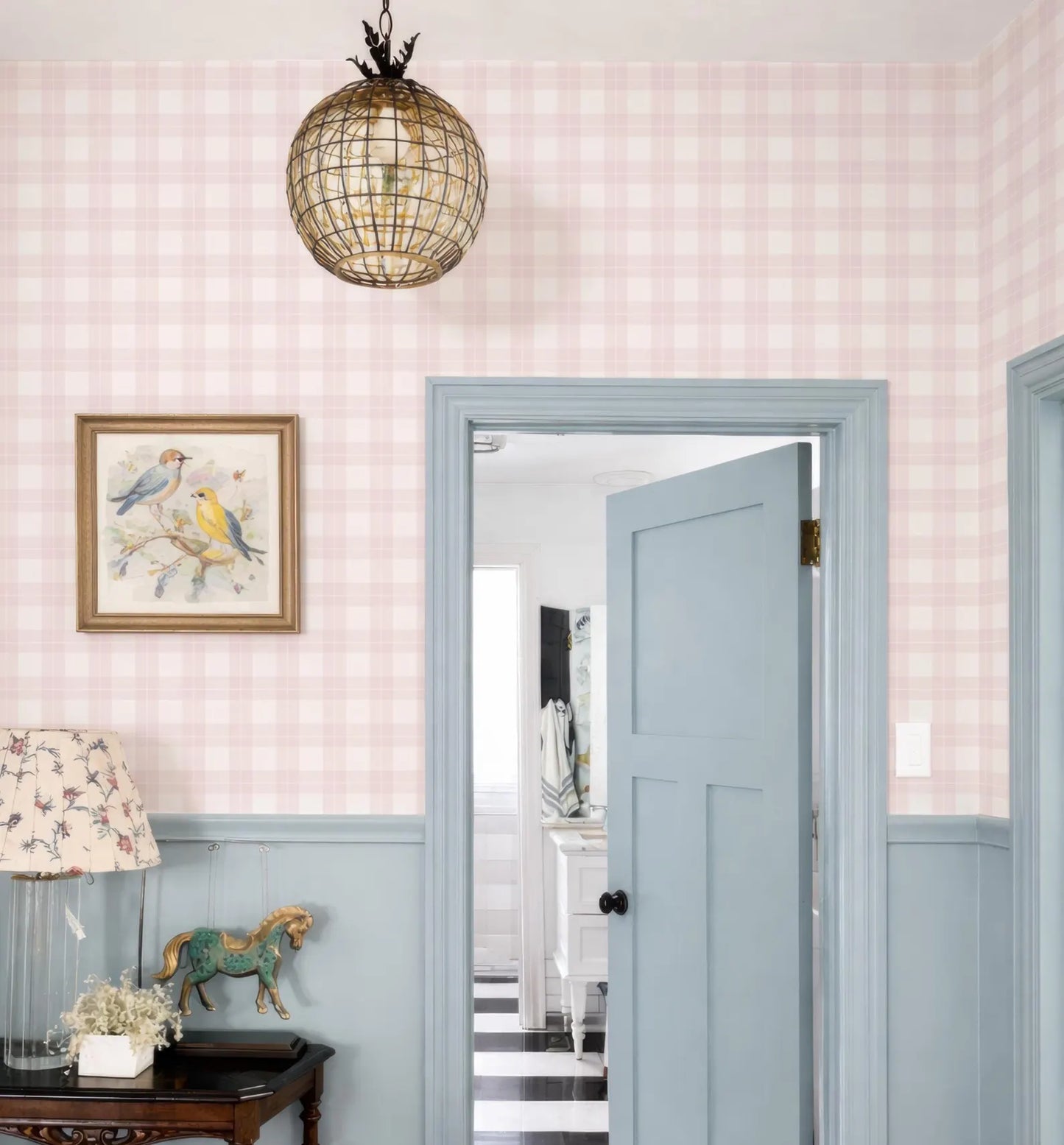 a room with a blue door, a pink and white checkered wallpaper, and a chandelier hanging above. There is a framed picture on the wall, and a small table with a lamp and a horse figurine. The door is open, and the hallway leads to another room.