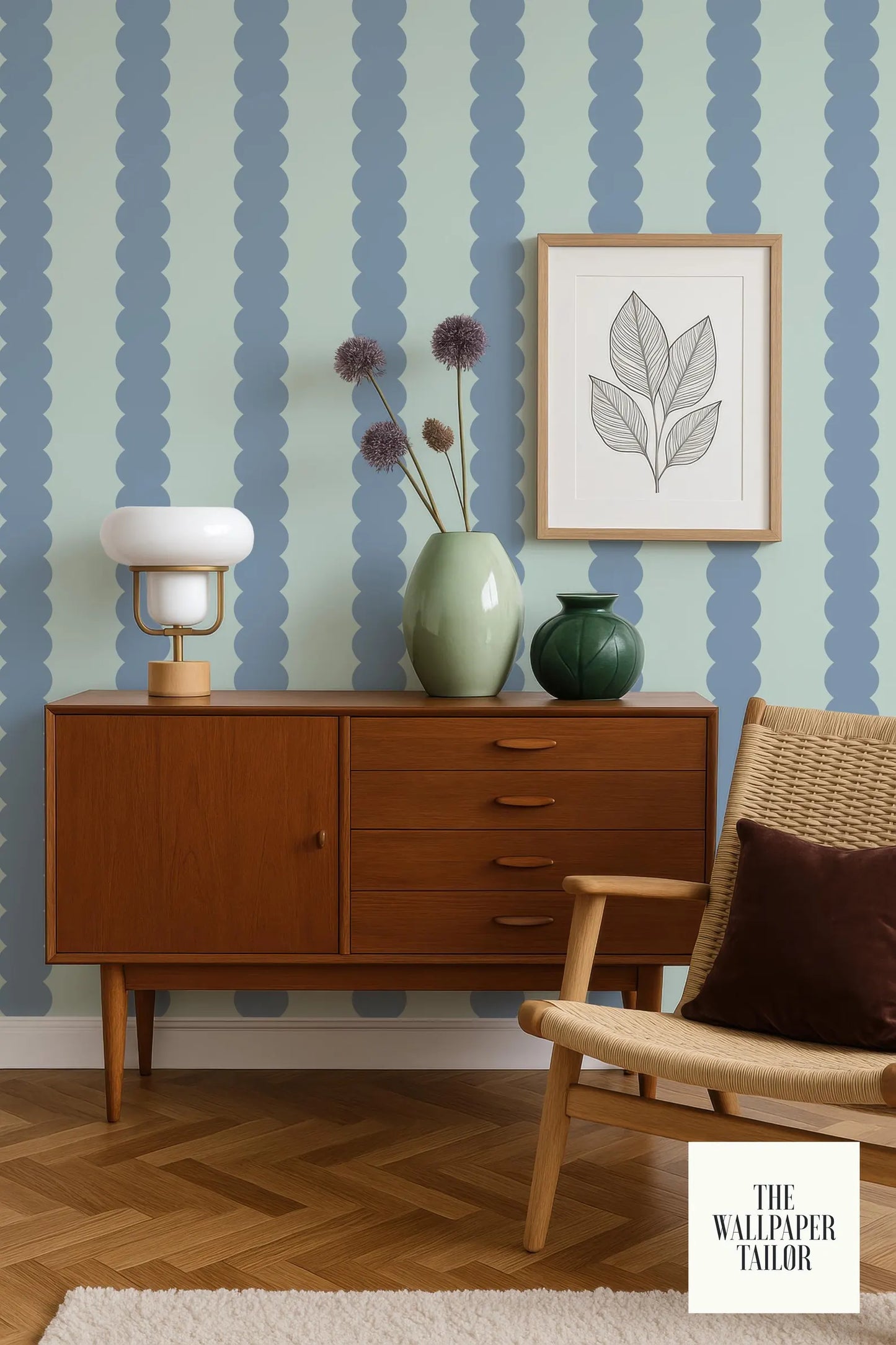 a modern living room with a wooden cabinet, a woven chair, and a framed artwork on the wall. The cabinet has a white lamp on top, and there are two vases on the cabinet. The chair has a brown pillow on it. The wall behind the cabinet has a blue and white striped pattern. The image also includes text that reads "The Wallpaper Tailor".