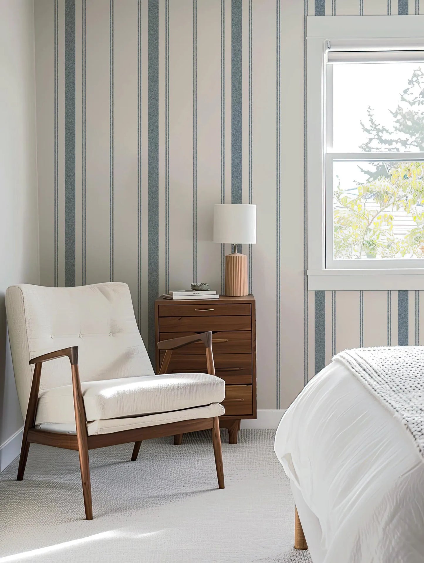 a cozy bedroom with a white armchair, a wooden side table with a lamp, and a window overlooking a tree outside.
