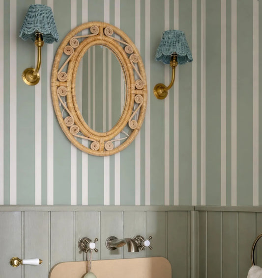 a bathroom with a light green and white striped wall, a round mirror with a woven frame, and two wall sconces with blue shades.