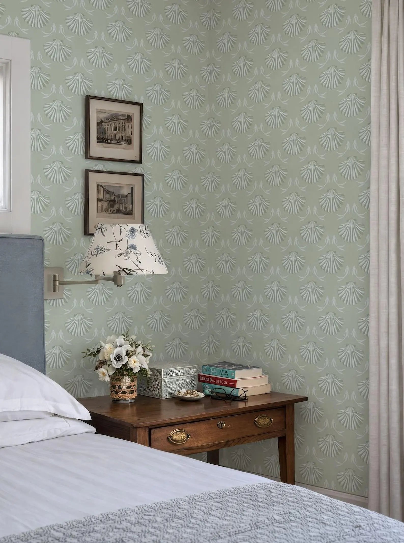 a cozy bedroom scene with a bed, a wooden nightstand, and a lamp. The walls are covered in a patterned wallpaper, and there are two framed pictures hanging above the bed. The nightstand has a vase of flowers and a stack of books on it.