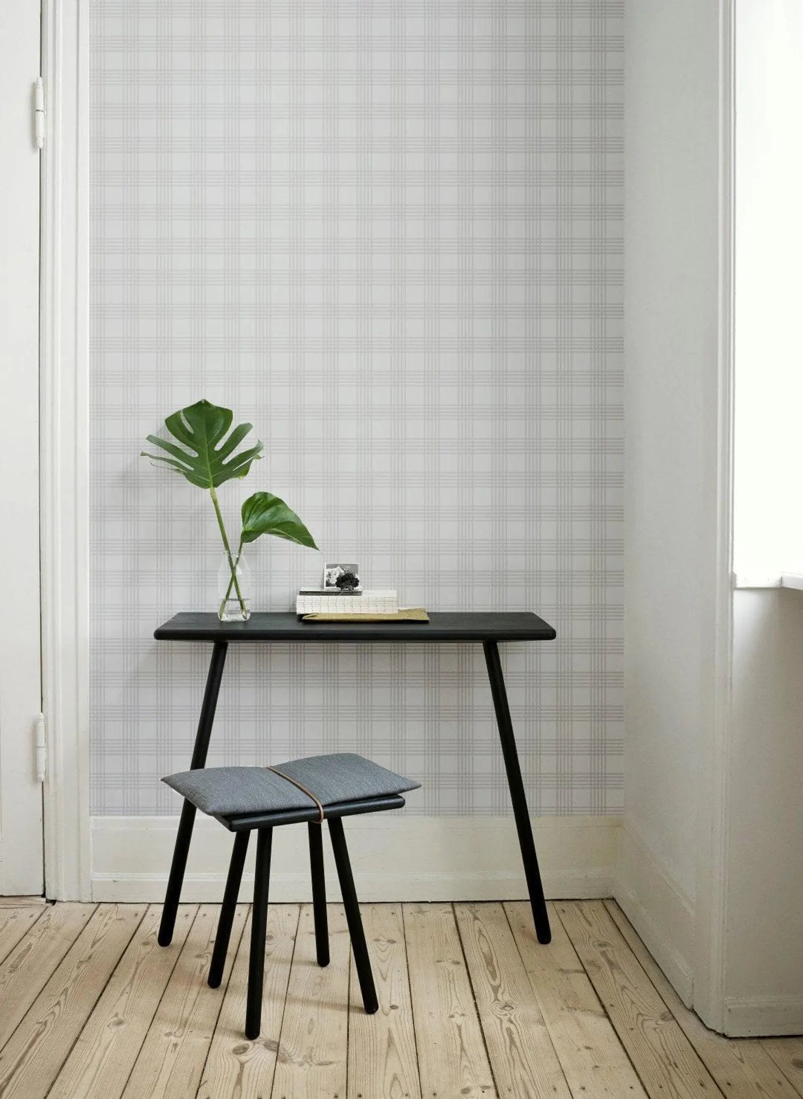 a minimalist interior design with a black desk, a small stool, and a potted plant. The desk has a book and a small vase on it, and the stool is positioned next to the desk. The room has a checkered pattern on the wall, and there is a window in the background.