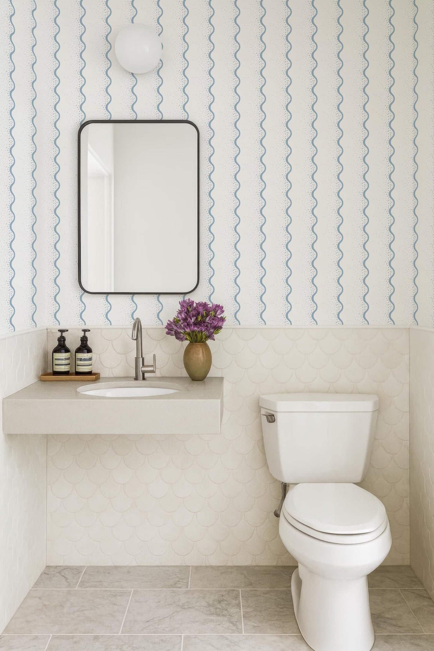 a bathroom with a white toilet, a sink with a mirror above it, and a vase of purple flowers on the counter. The walls are covered in blue and white patterned wallpaper, and the floor is tiled in white.