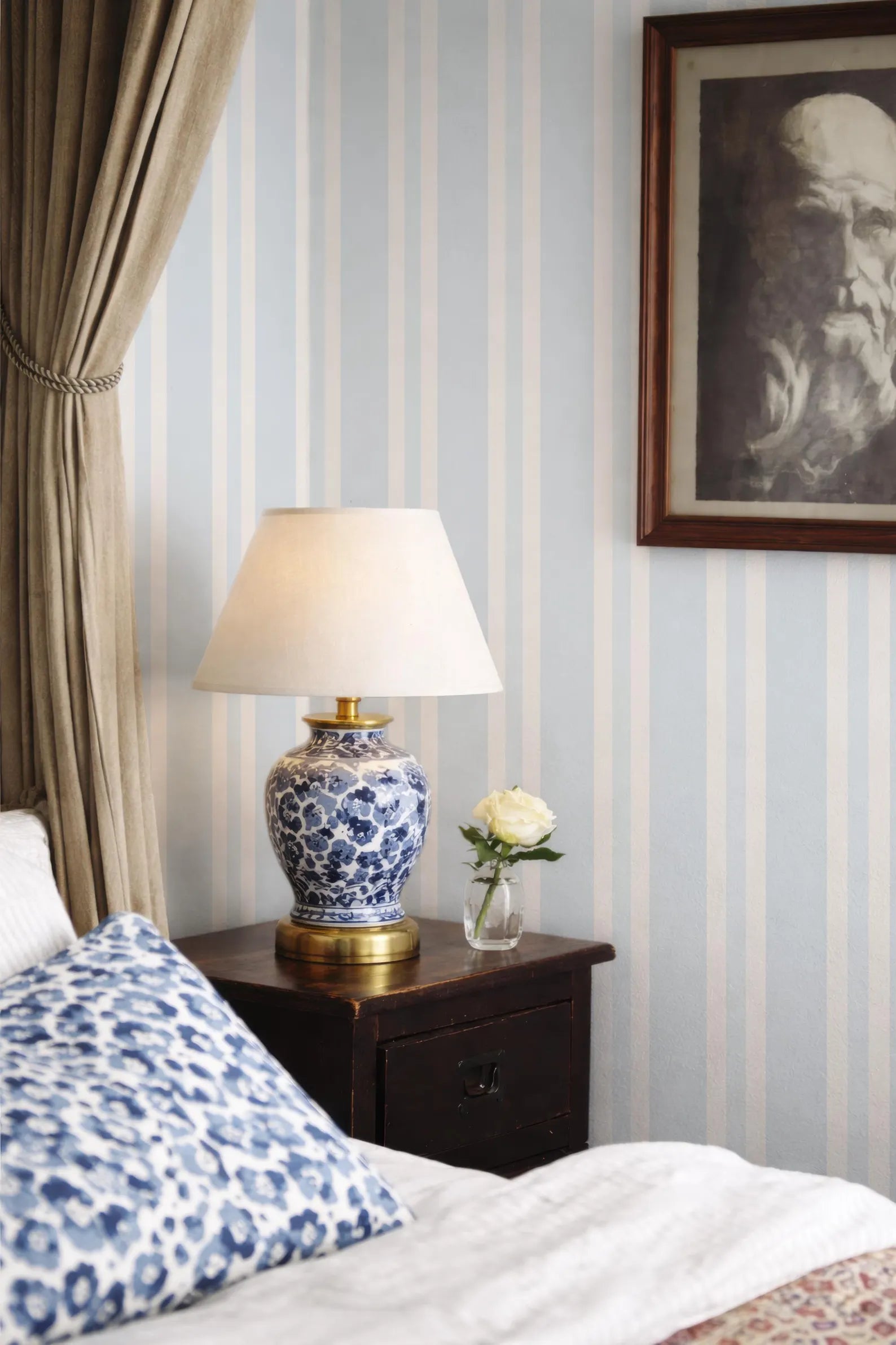 a bedroom scene with a blue and white patterned pillow, a wooden nightstand with a lamp and a vase of flowers, and a framed picture on the wall.