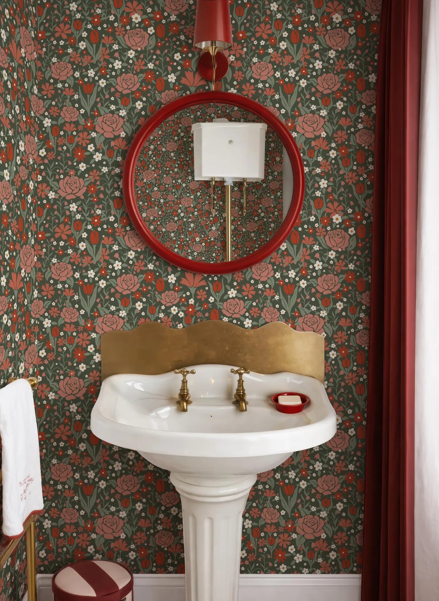 a bathroom with a white pedestal sink, a red circular mirror, and a floral wallpaper.