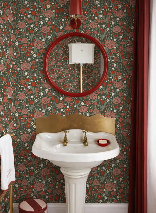 a bathroom with a white pedestal sink, a red circular mirror, and a floral wallpaper.