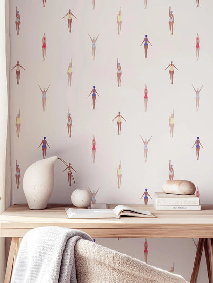 a wallpapered wall with a repeating pattern of stylized female figures in various poses, creating a vibrant and dynamic visual effect. In the foreground, there is a wooden desk with a white vase, a book, and a small white figurine.