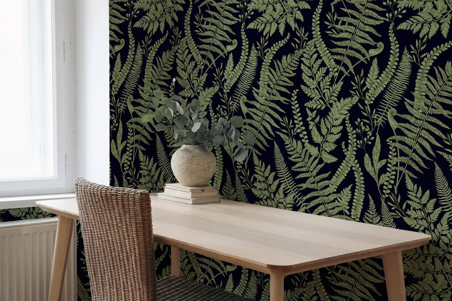 a wooden table with a vase of greenery on top, set against a wall with a dark green fern pattern. The table is positioned near a window, and there is a wicker chair visible in the foreground.