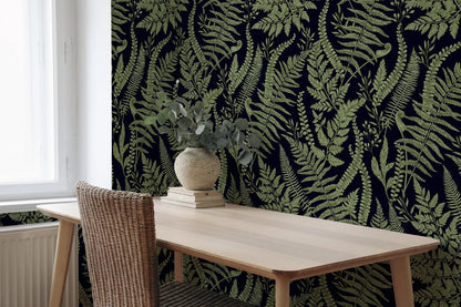 a wooden table with a vase of greenery on top, set against a wall with a dark green fern pattern. The table is positioned near a window, and there is a wicker chair visible in the foreground.