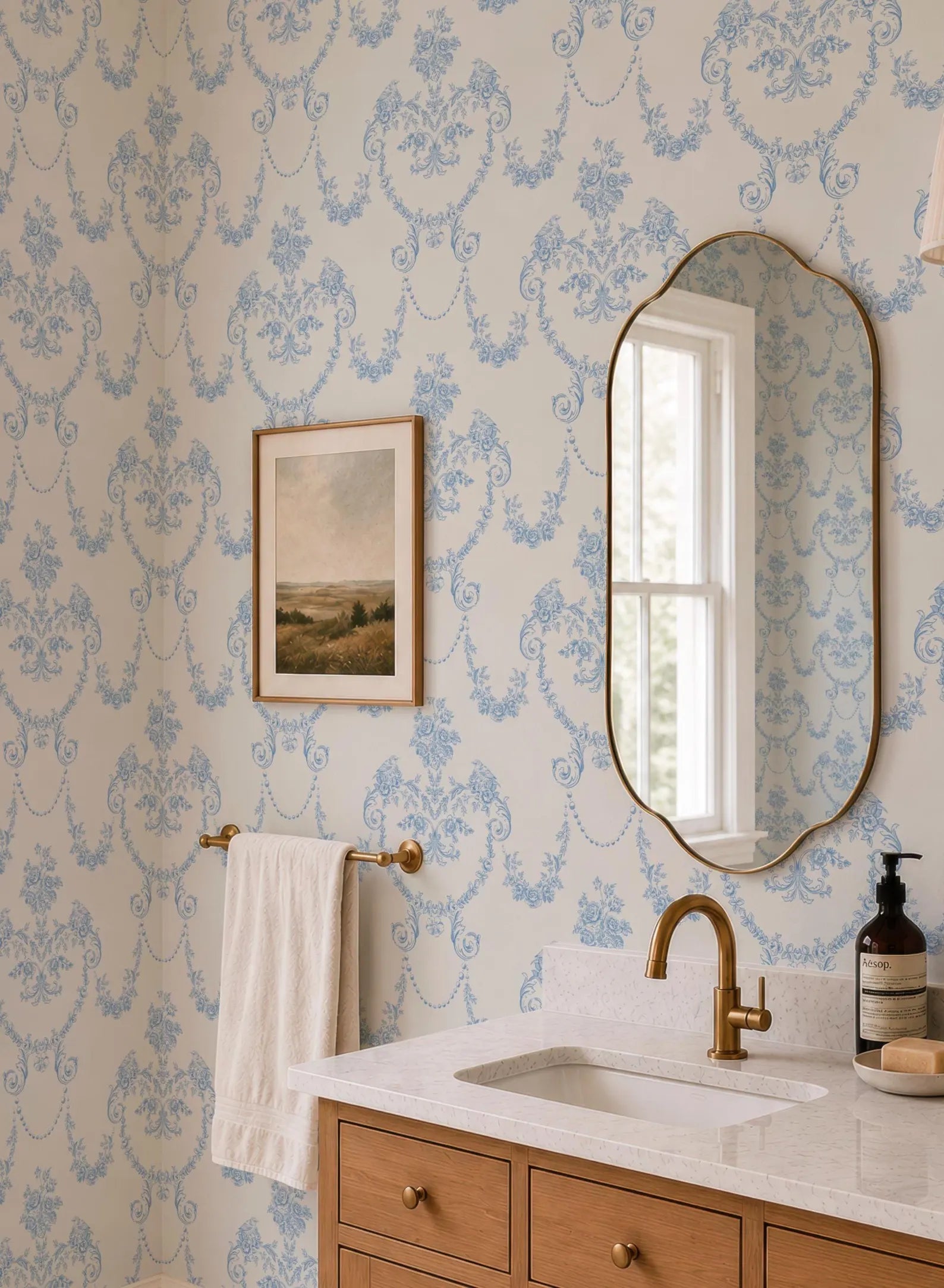 a bathroom with a white sink, a mirror, and a towel rack. The walls are covered in blue floral wallpaper, and there is a painting hanging above the mirror.