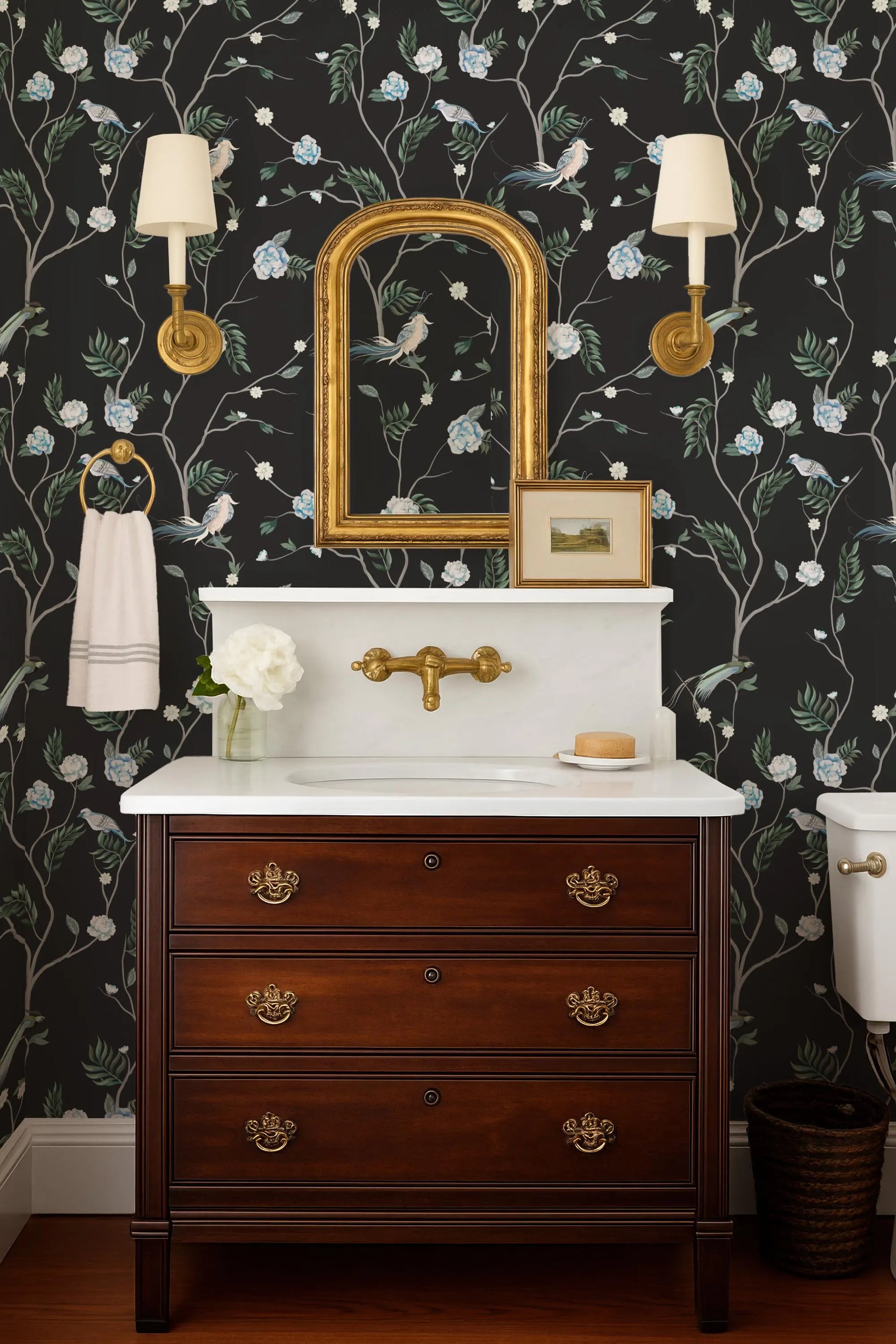 a bathroom with a white sink, a wooden dresser, and a mirror above the sink. The walls are decorated with a floral wallpaper, and there are two wall sconces on either side of the mirror.