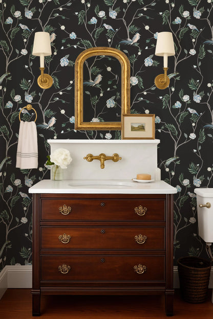 a bathroom with a white sink, a wooden dresser, and a mirror above the sink. The walls are decorated with a floral wallpaper, and there are two wall sconces on either side of the mirror.