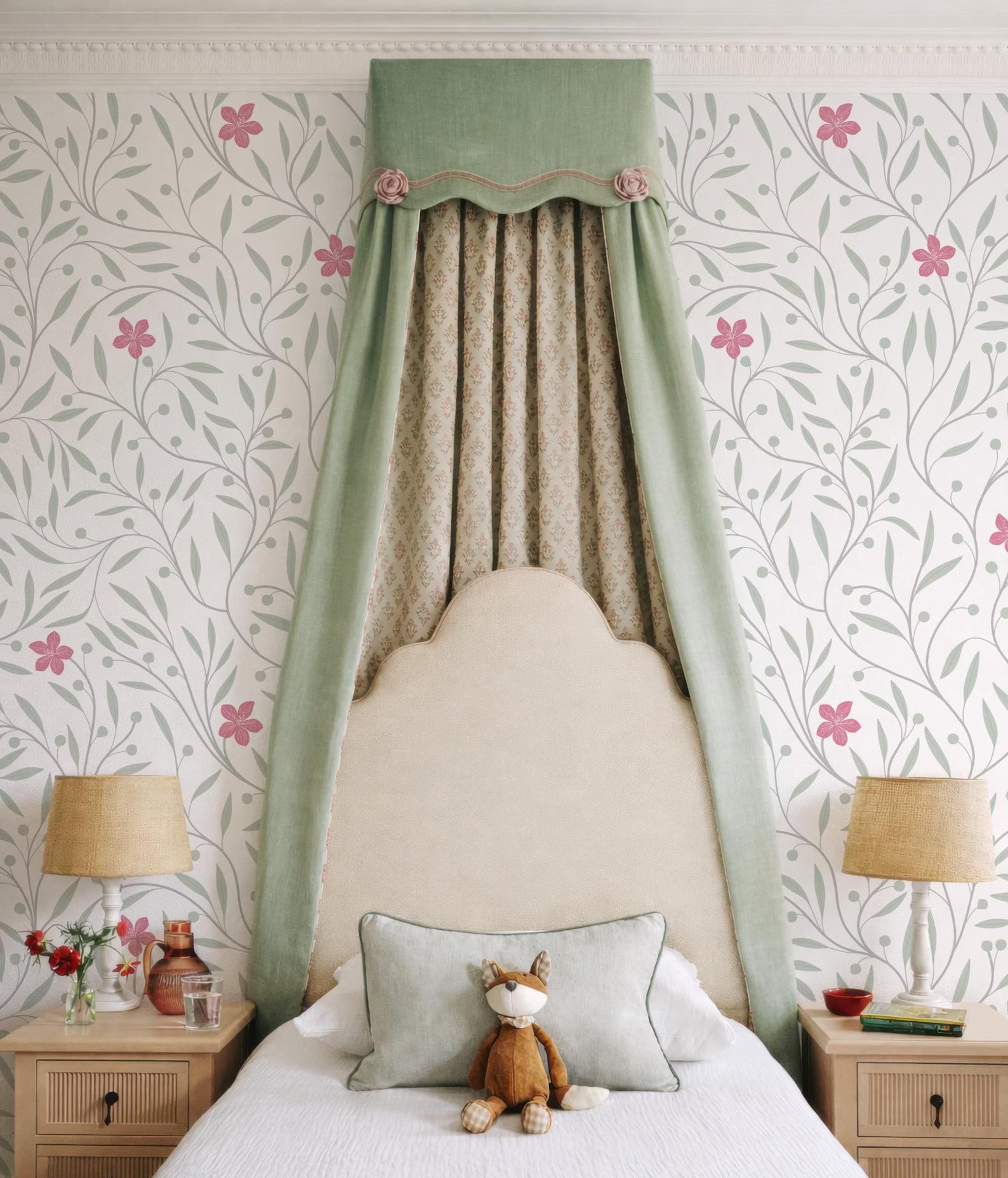 a cozy bedroom with a white bed, a green curtain, and a lamp on a nightstand. A stuffed animal is placed on the bed, and the walls are decorated with a floral pattern.
