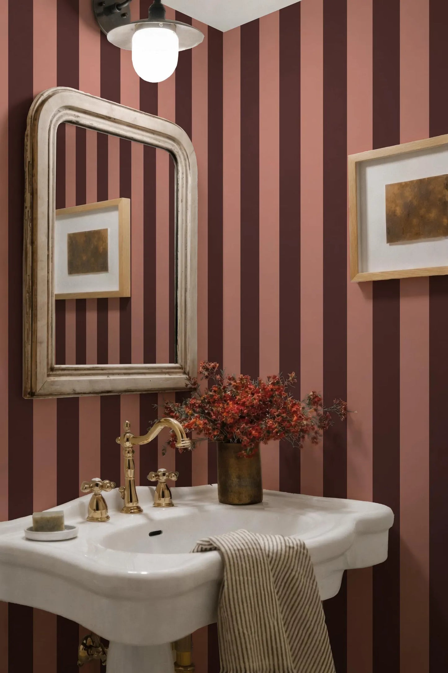 a bathroom with a white pedestal sink, a gold faucet, and a framed picture on the wall. The walls are striped in shades of pink and burgundy, and there is a vase of red flowers on the sink.