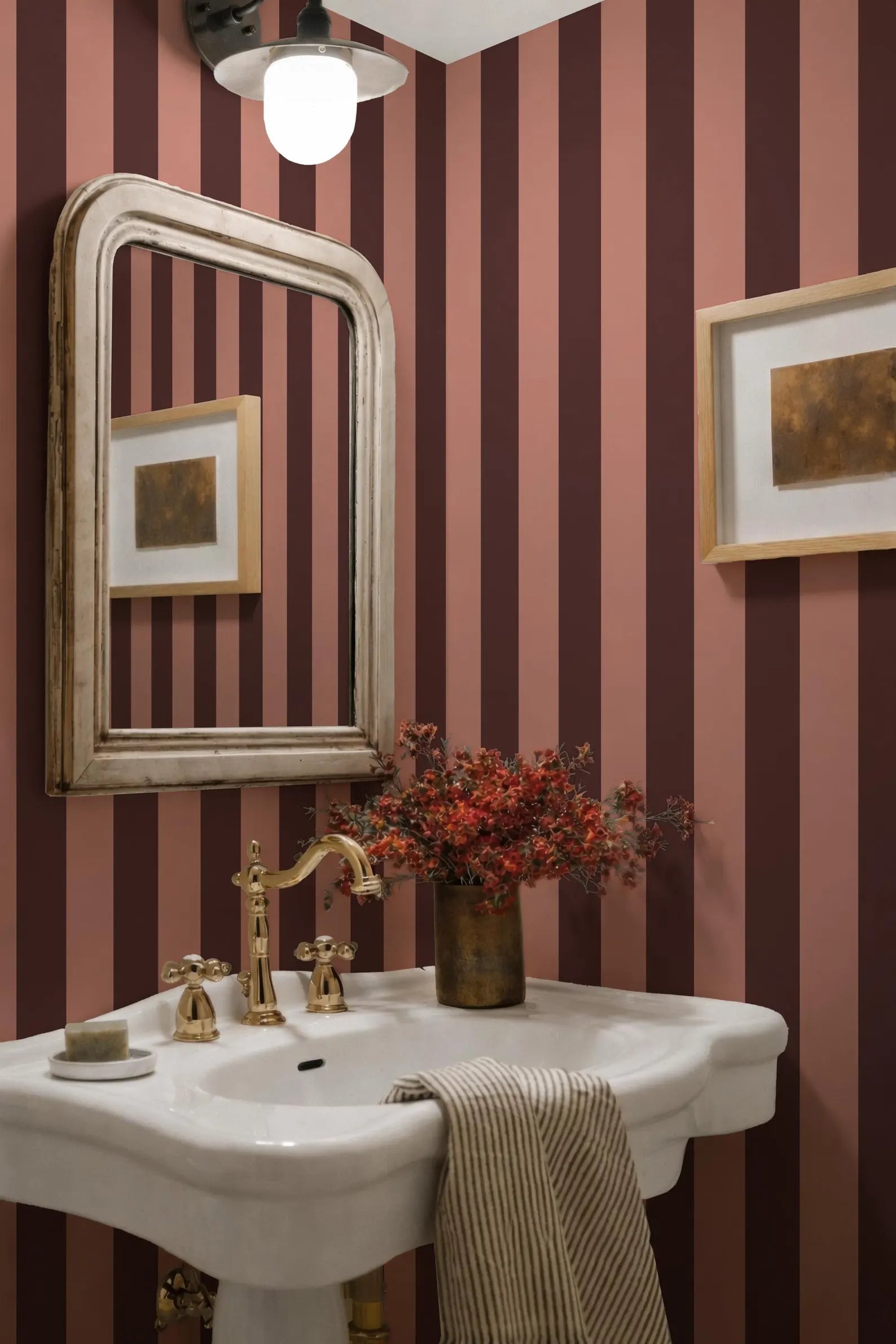 a bathroom with a white pedestal sink, a gold faucet, and a framed picture on the wall. The walls are striped in shades of pink and burgundy, and there is a vase of red flowers on the sink.