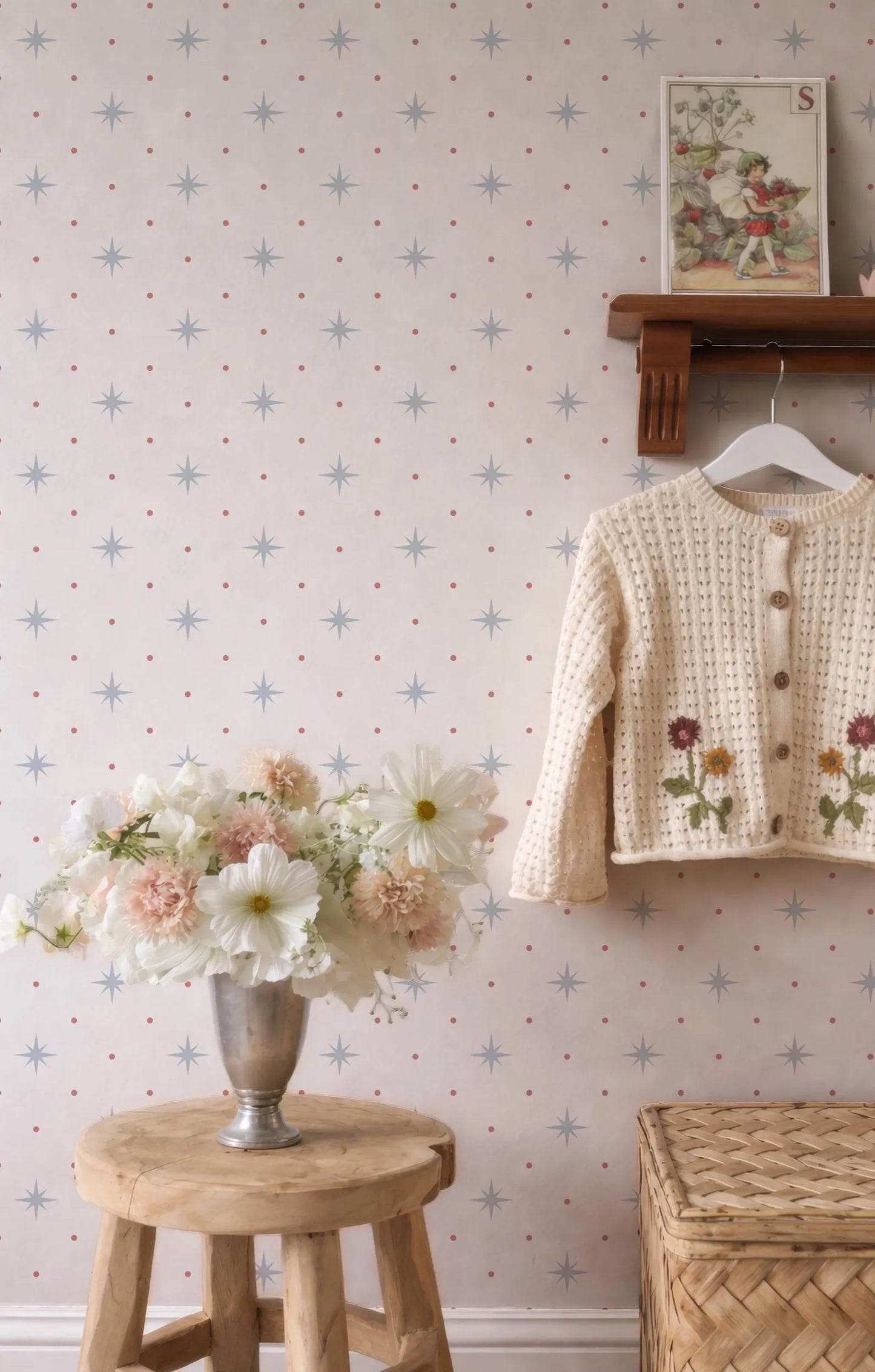 a cozy and charming interior scene with a wooden stool holding a vase filled with white and pink flowers, a wooden shelf displaying a white sweater with floral embroidery, and a wooden shelf with a framed picture. The wall behind these items is decorated with a pattern of blue and white stars.